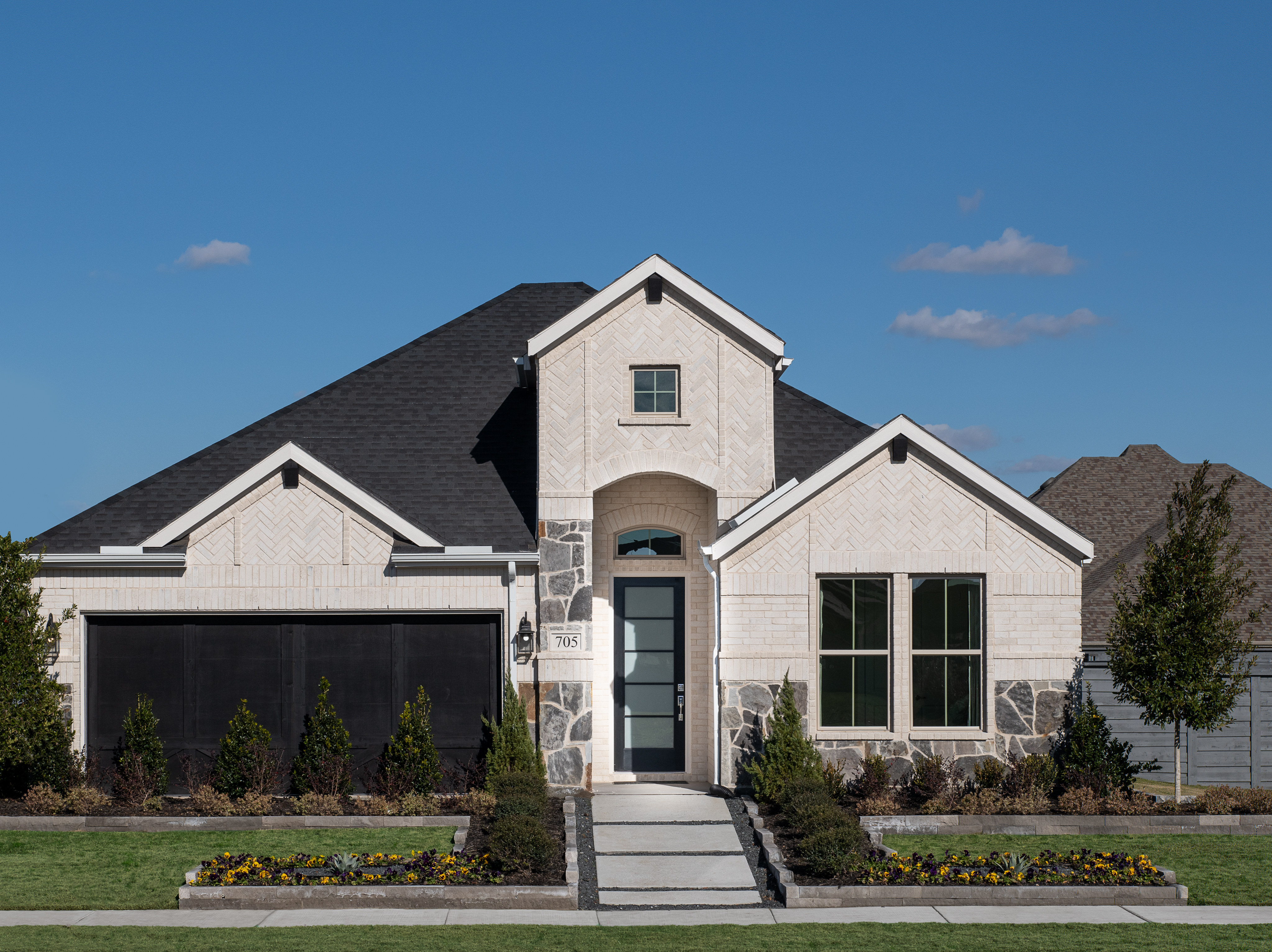 A two-story house with a gabled roof, stone and siding exterior, and a landscaped front yard with shrubs and flowers, set against a clear blue sky with a few clouds.