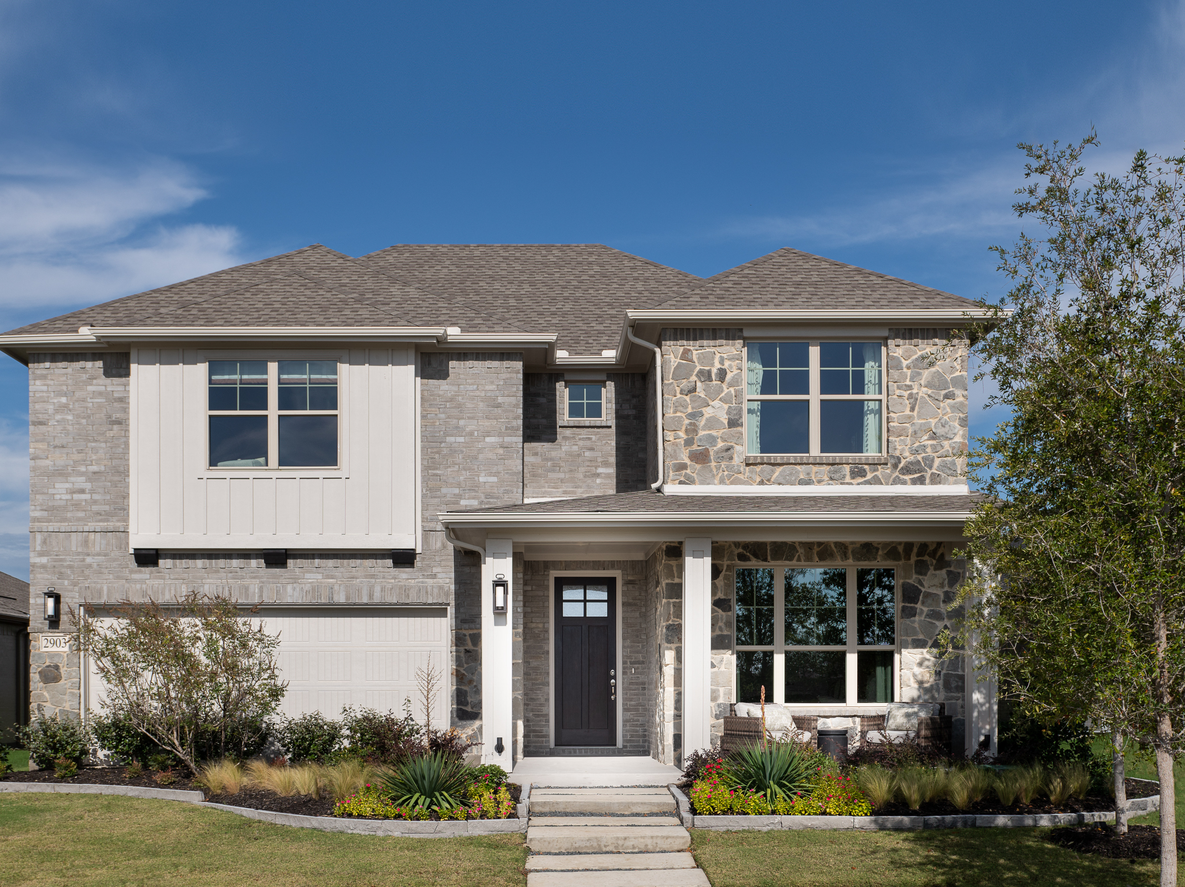 A two-story residential house with a stone and stucco exterior, surrounded by a well-manicured lawn and landscaping, set against a clear blue sky with scattered clouds.