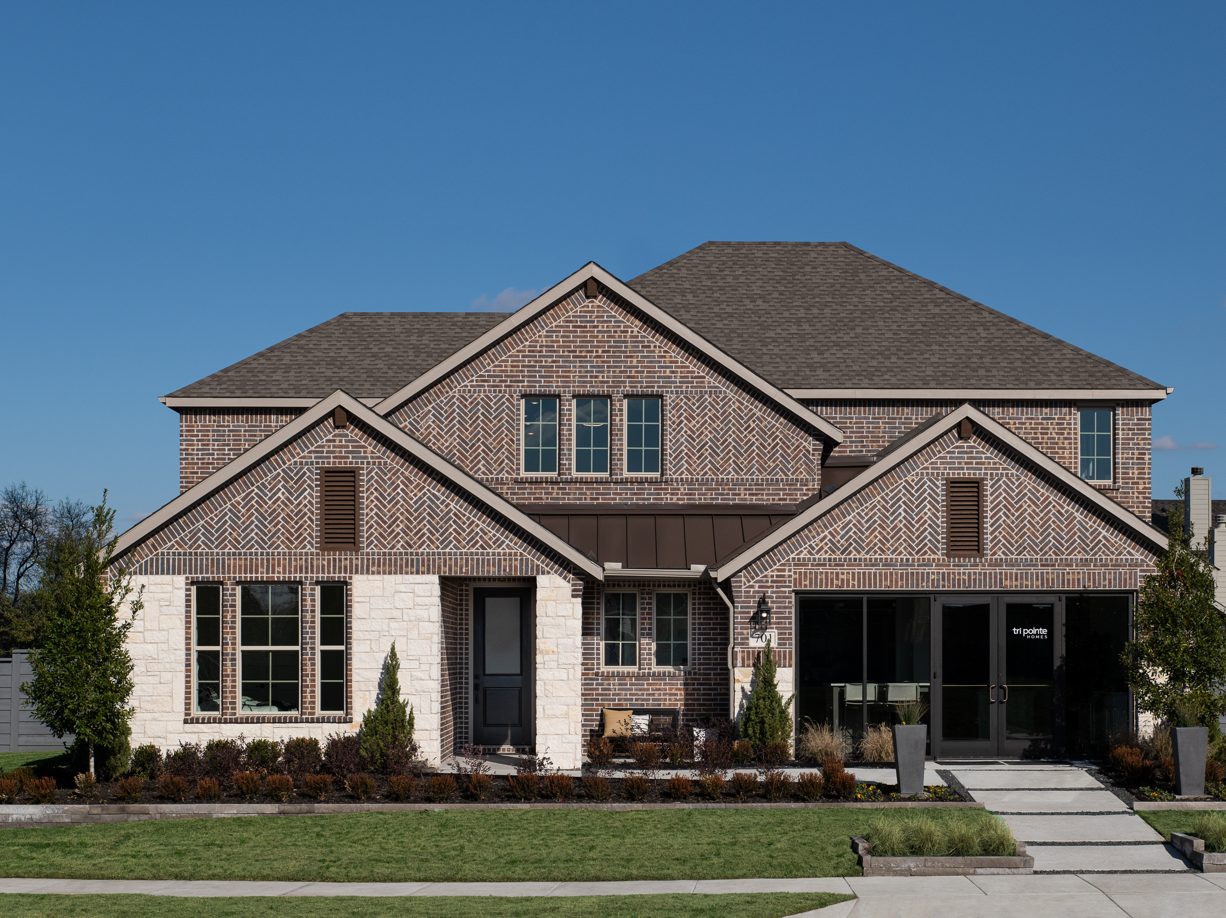 A two-story brick house with a gabled roof, surrounded by landscaped gardens and a paved driveway, set against a clear blue sky.