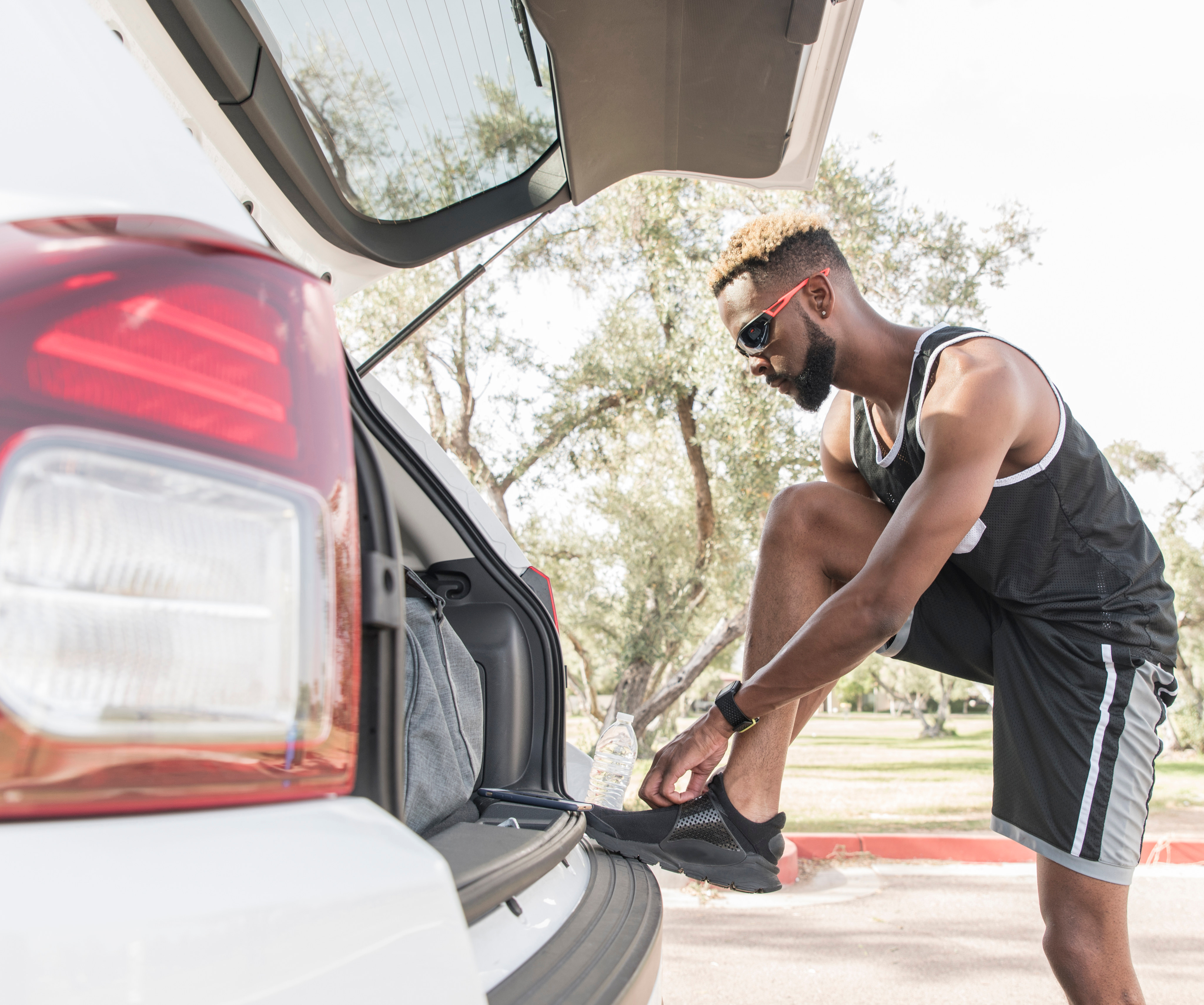 A man in athletic clothing is leaning into the open trunk of a red car, surrounded by a grassy outdoor setting.