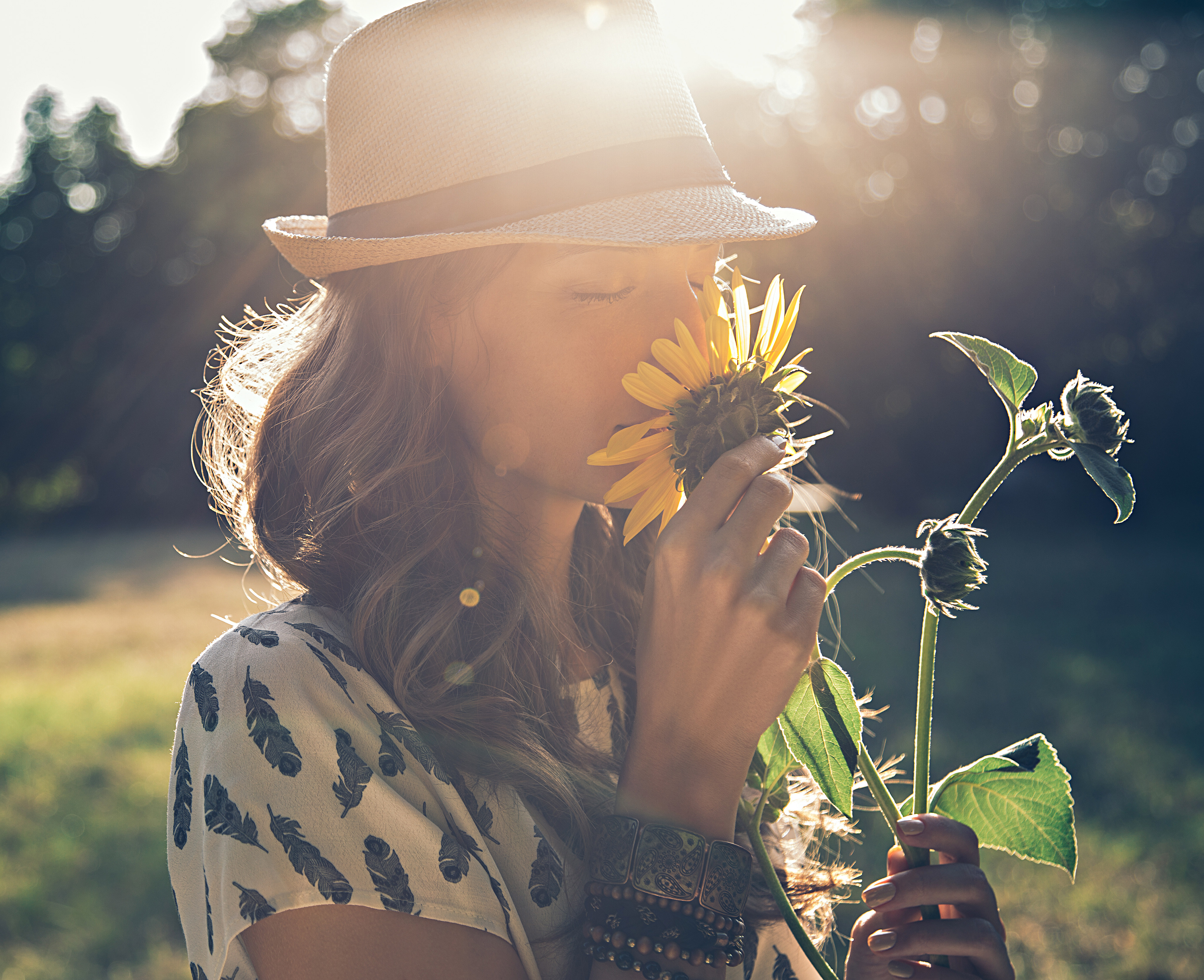 A person wearing a hat and a polka dot shirt stands in a field, holding a sunflower and surrounded by lush greenery and sunlight.