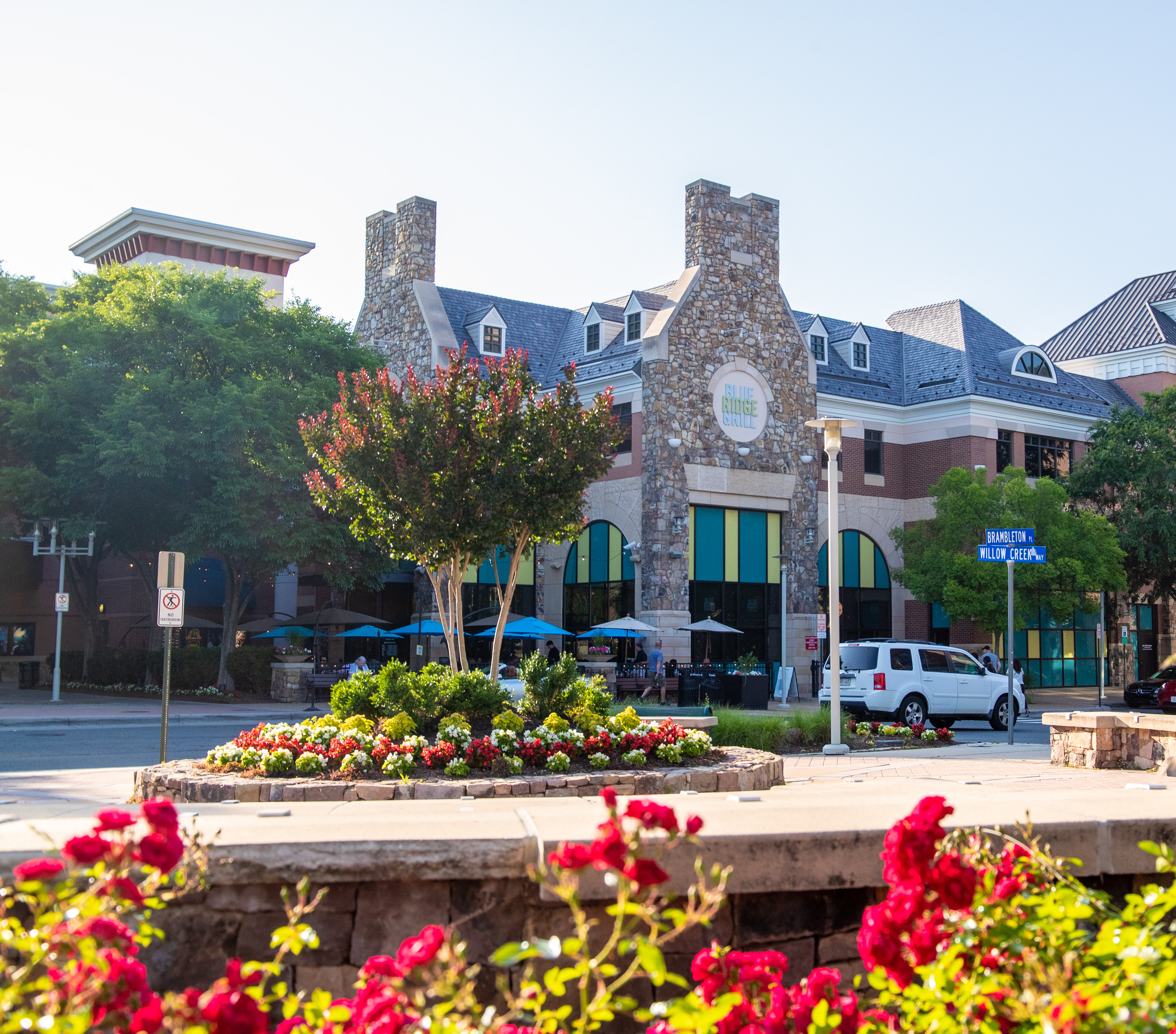 A vibrant and colorful urban scene with a stone fountain surrounded by blooming flowers in the foreground, and a collection of distinctive stone and brick buildings in the background.