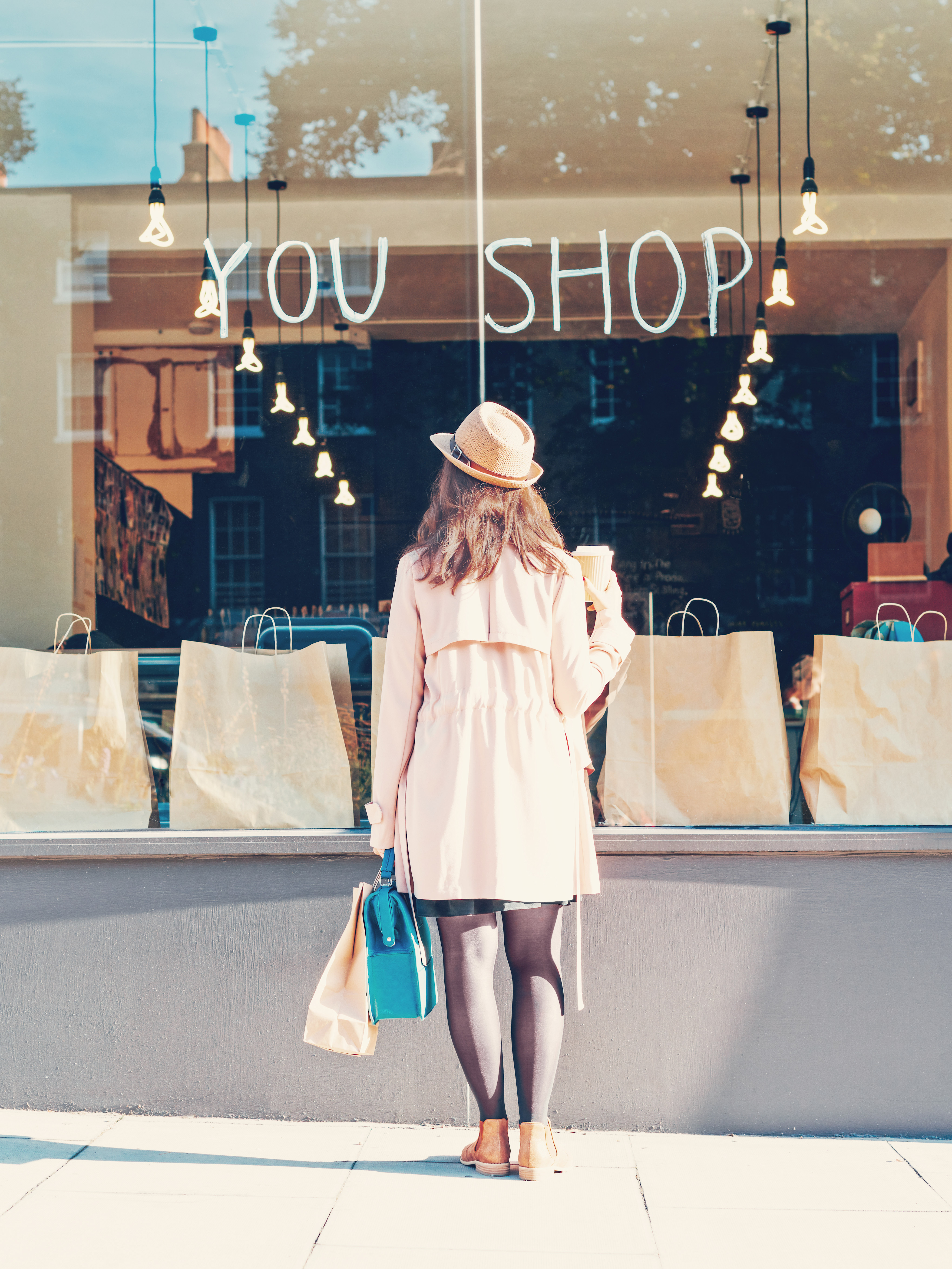 A woman in a light-colored coat and hat stands in front of a storefront with the words "YOU SHOP" displayed on the window, surrounded by shopping bags and a warm, inviting atmosphere.