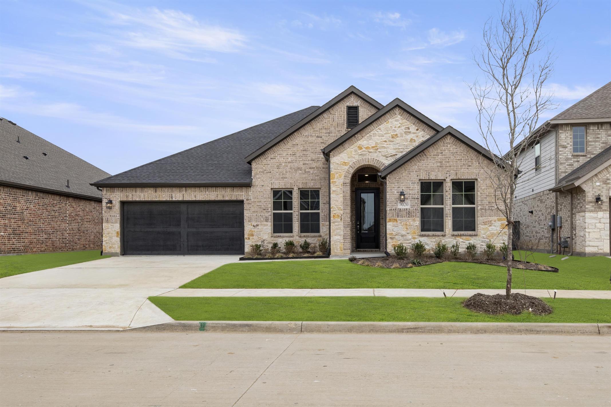 A two-story brick and stone house with a garage, surrounded by a well-manicured lawn and landscaping, set against a clear blue sky with scattered clouds.