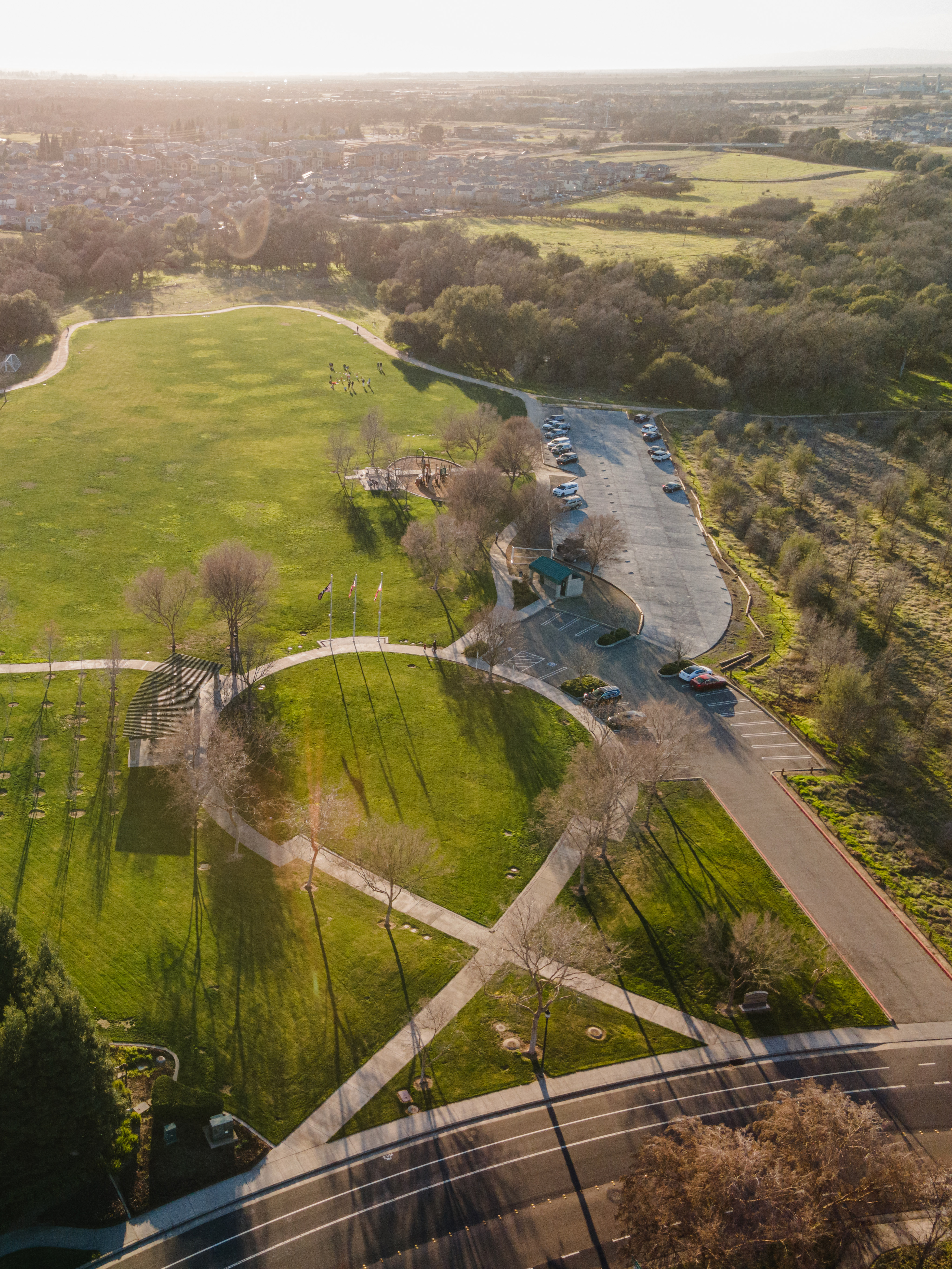 A lush, green park with winding paths and buildings in the distance, surrounded by a rural landscape with fields and forests.
