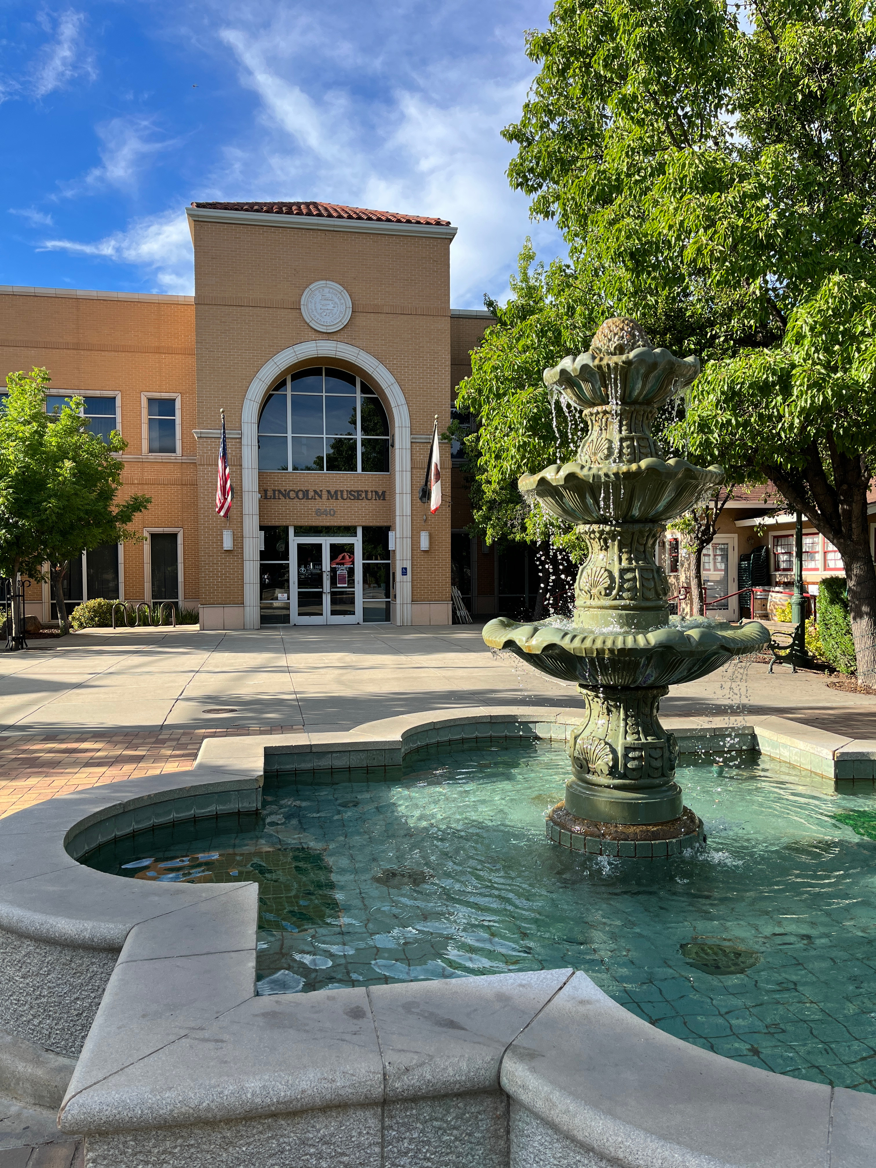 A decorative fountain sits in the foreground, surrounded by a paved plaza, with a two-story building with an arched entryway in the background, framed by lush greenery.