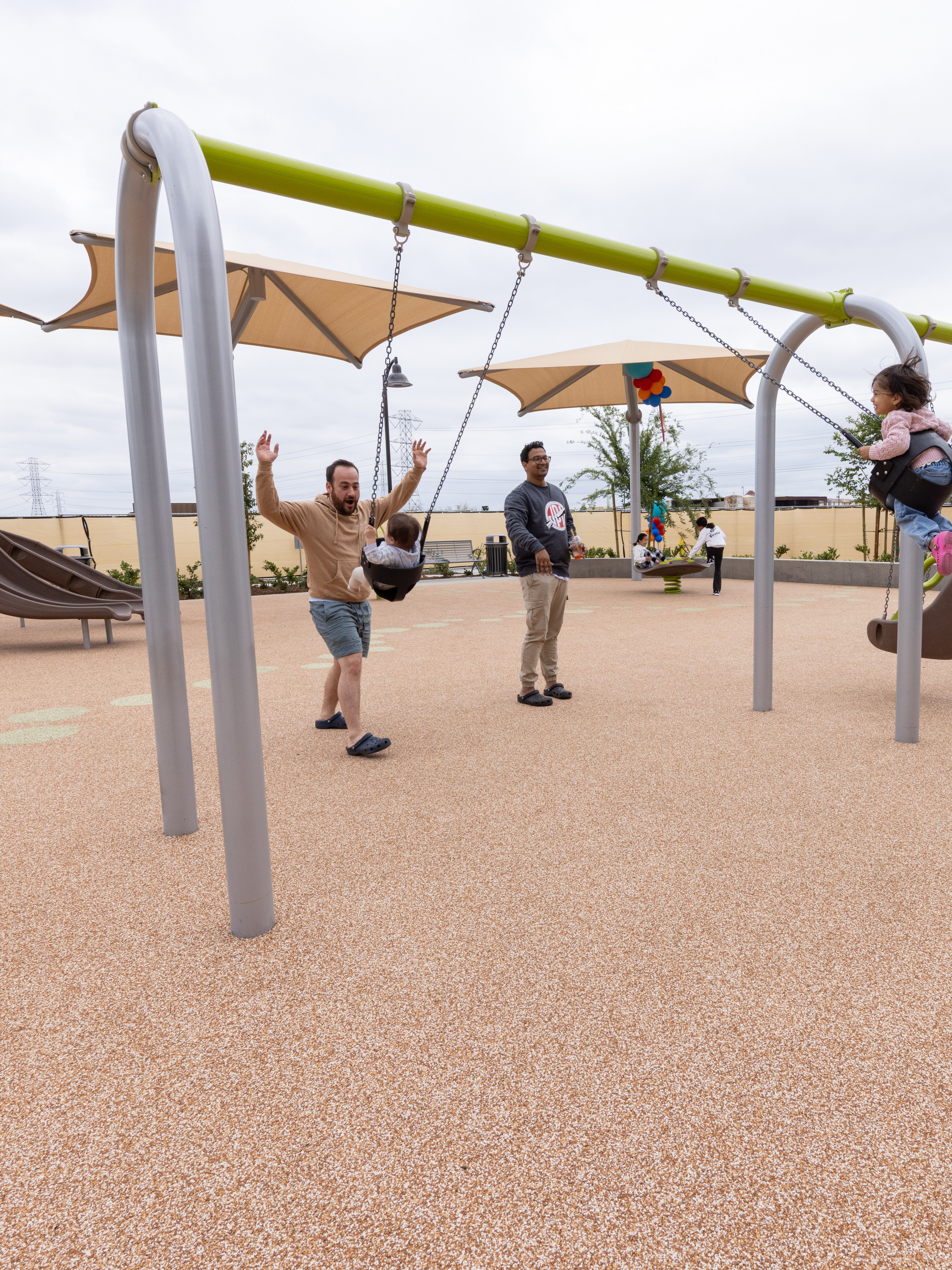 A modern outdoor playground with various colorful equipment and structures, surrounded by residential buildings and shaded areas.