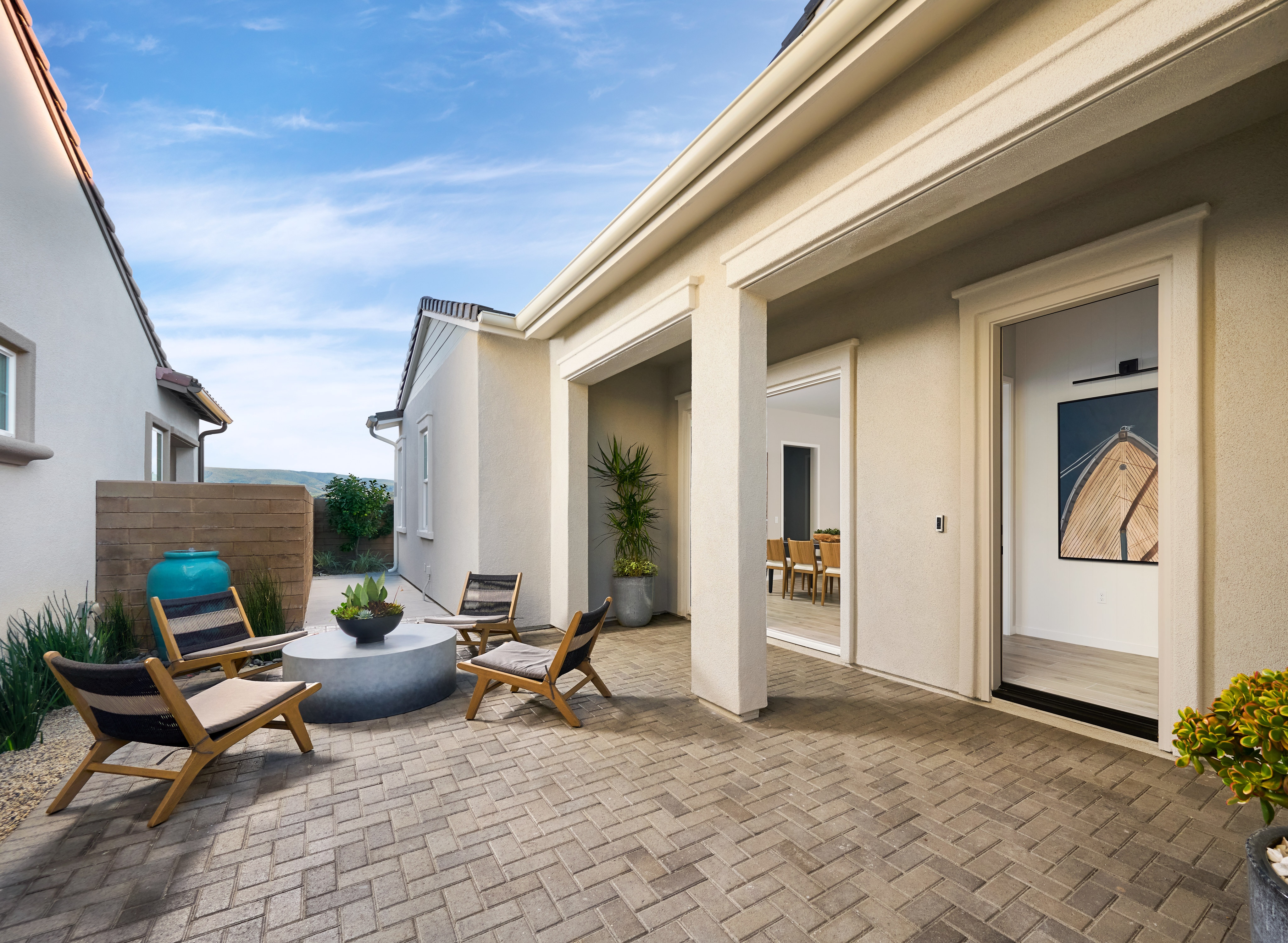 A cozy outdoor patio with wooden furniture, potted plants, and a brick-paved floor, surrounded by a modern, beige-colored house with large windows and an inviting entryway.