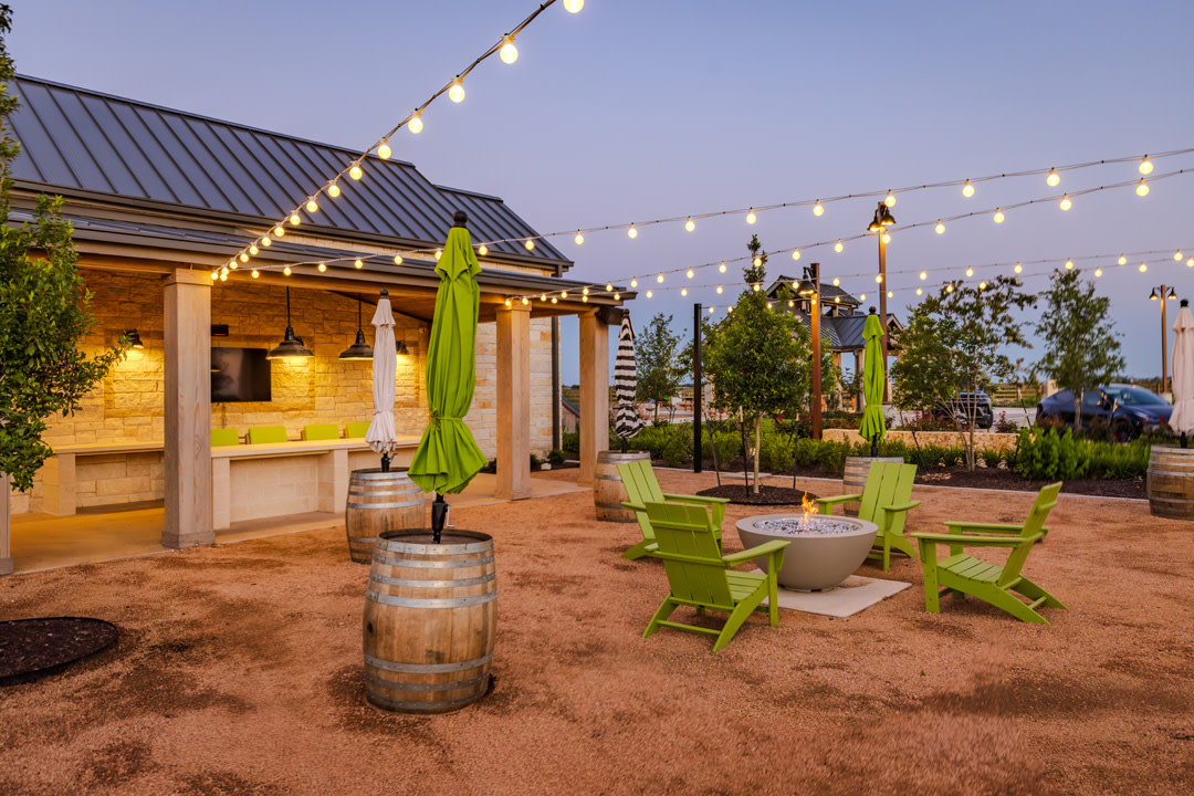 In the foreground, lime green Adirondack chairs surround a fire pit on a gravel patio with wine barrels and umbrellas, while the background features a covered outdoor kitchen area with a bar and string lights illuminating the twilight sky.