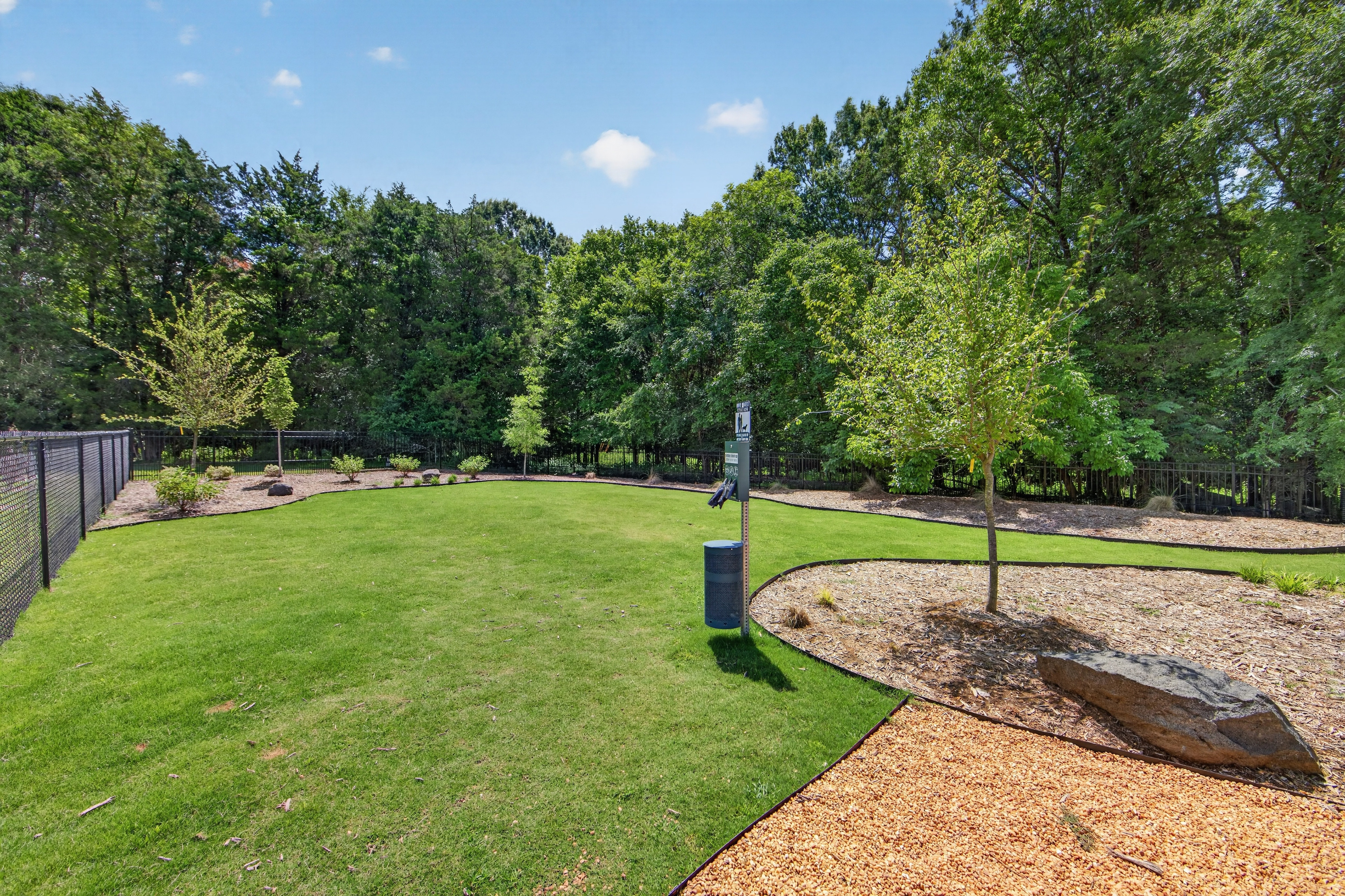 A lush green dog park with a waste station in the foreground is surrounded by a dense forest in the background.