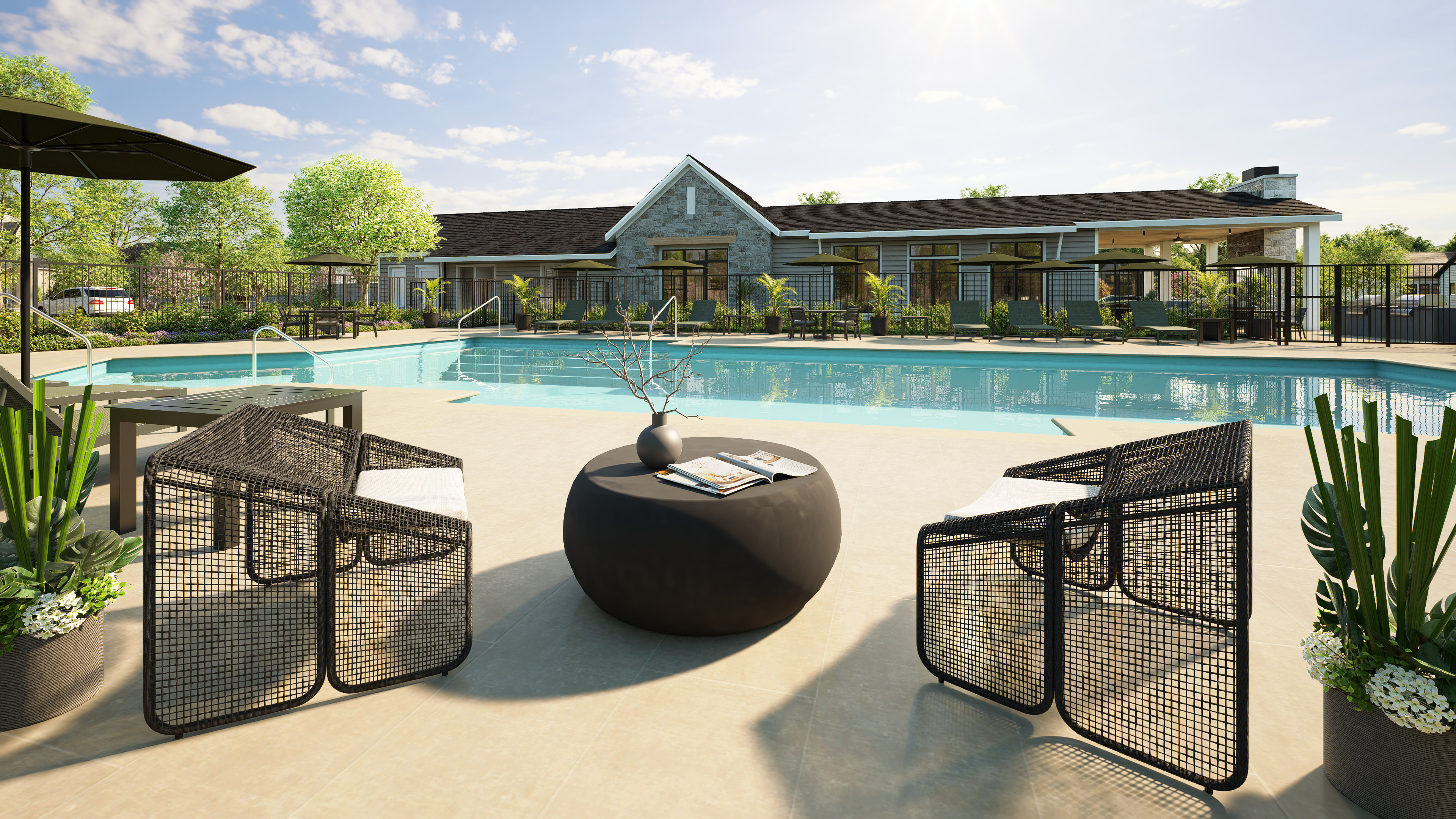 Two modern chairs and a round table with a vase and book sit on a patio in the foreground, while a large swimming pool and a stone-clad building with an overhang are in the background.