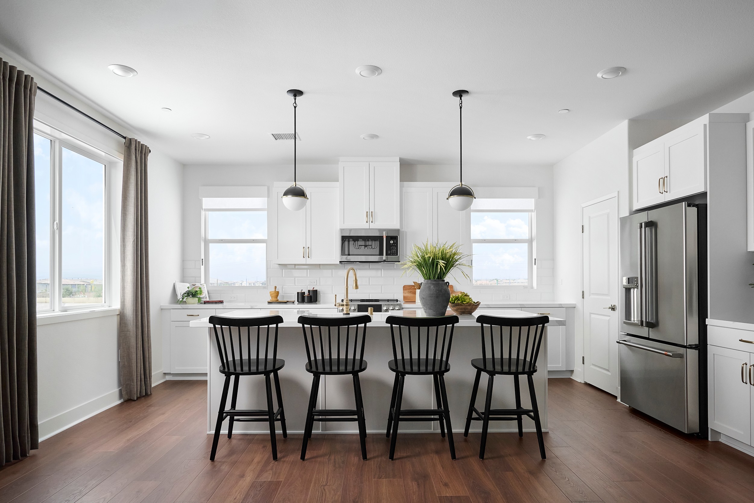 Four black bar stools sit at a white kitchen island with a bouquet of flowers, while a stainless steel refrigerator and white cabinets fill the background.