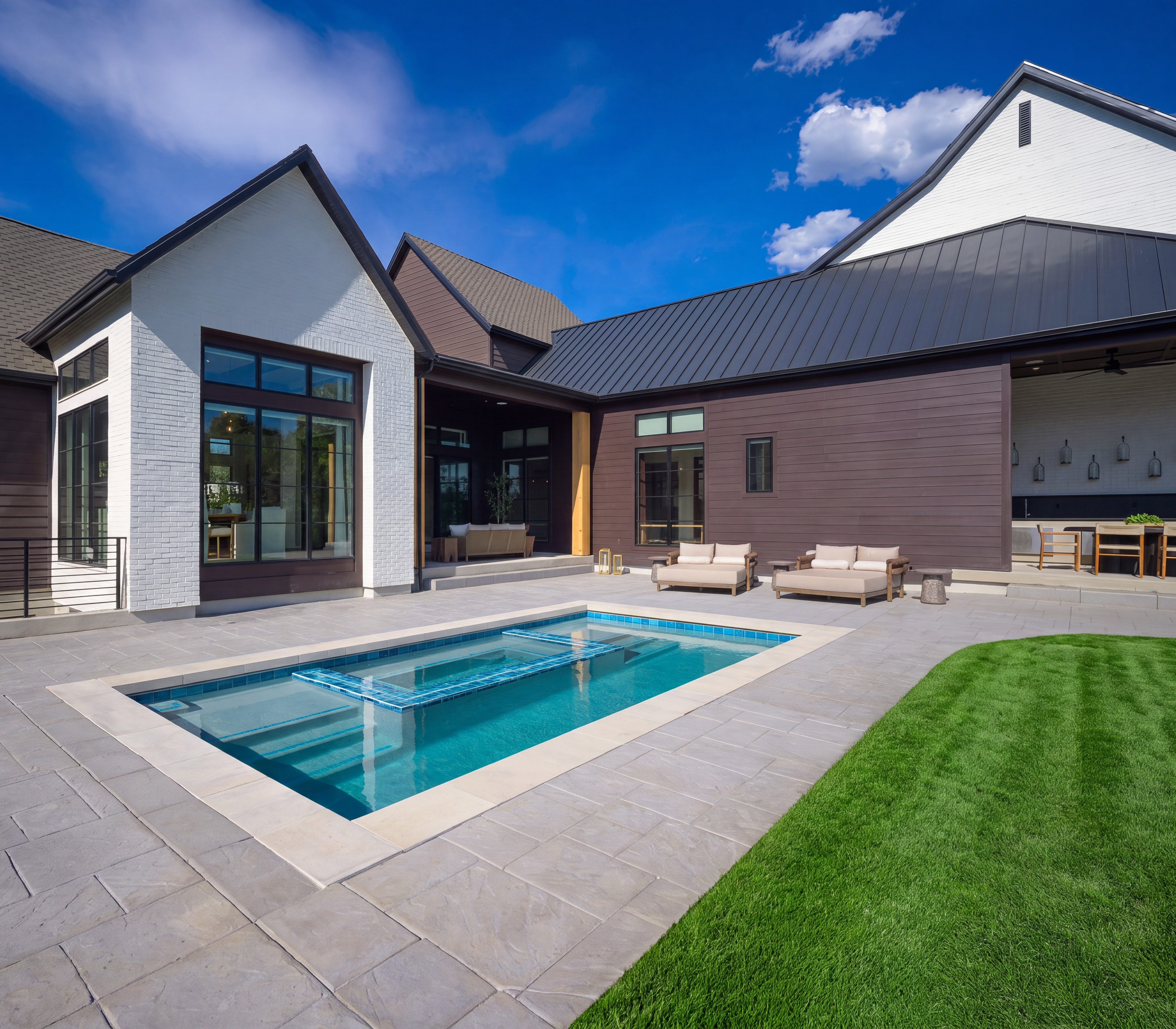 A rectangular swimming pool with a tiled insert sits on a paved patio in the foreground, while a modern house with dark siding and white accents, along with a manicured lawn, forms the background under a bright blue sky.