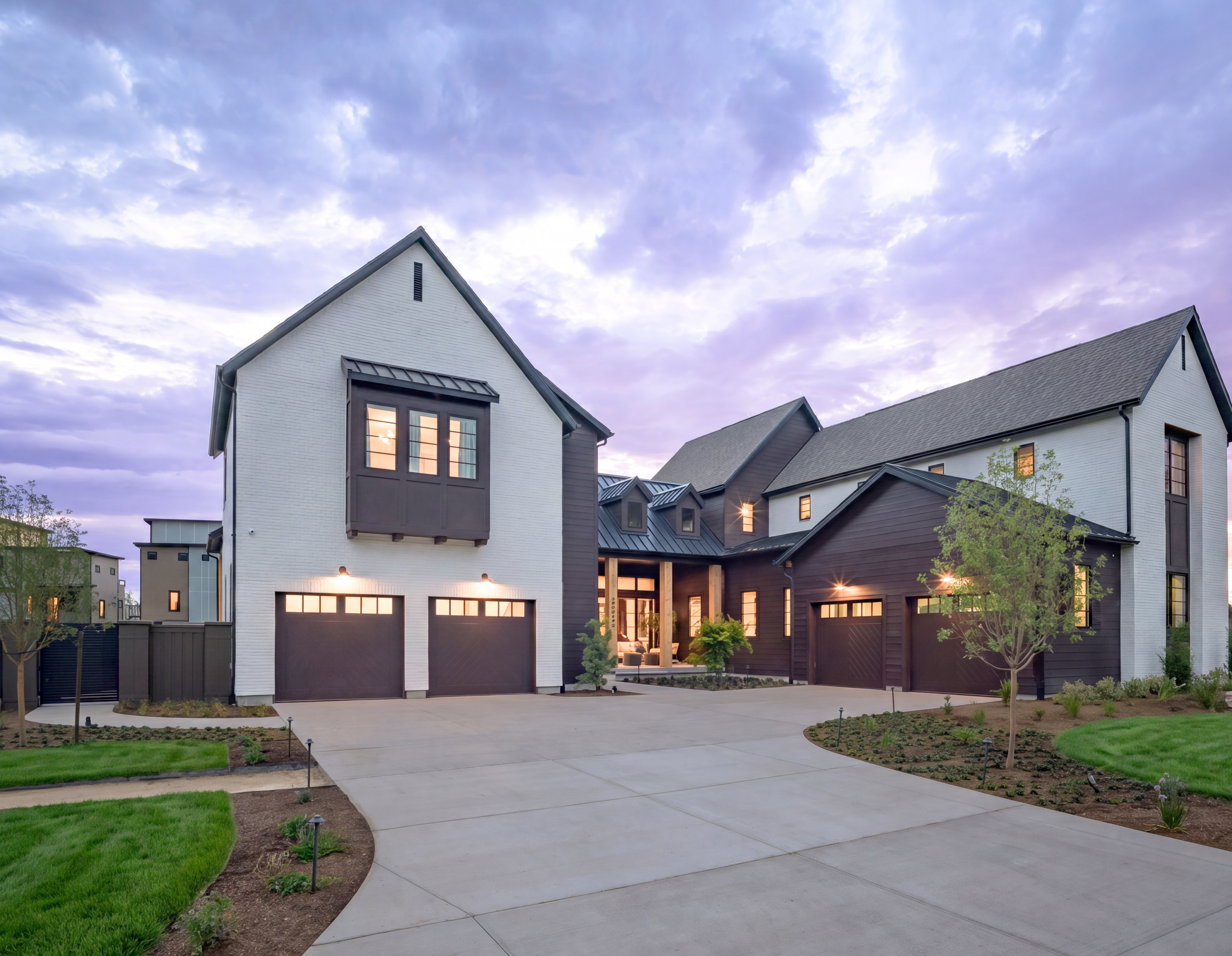A modern white and dark wood house with a wide driveway in the foreground sits under a dramatic purple and pink sky.