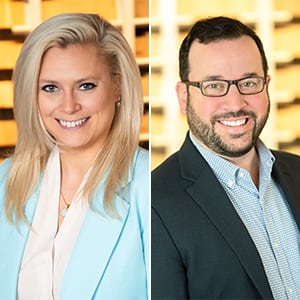 The image shows a smiling woman with long blonde hair wearing a light blue top, and a smiling man with glasses and a beard wearing a dark suit, both standing in front of a wooden background.