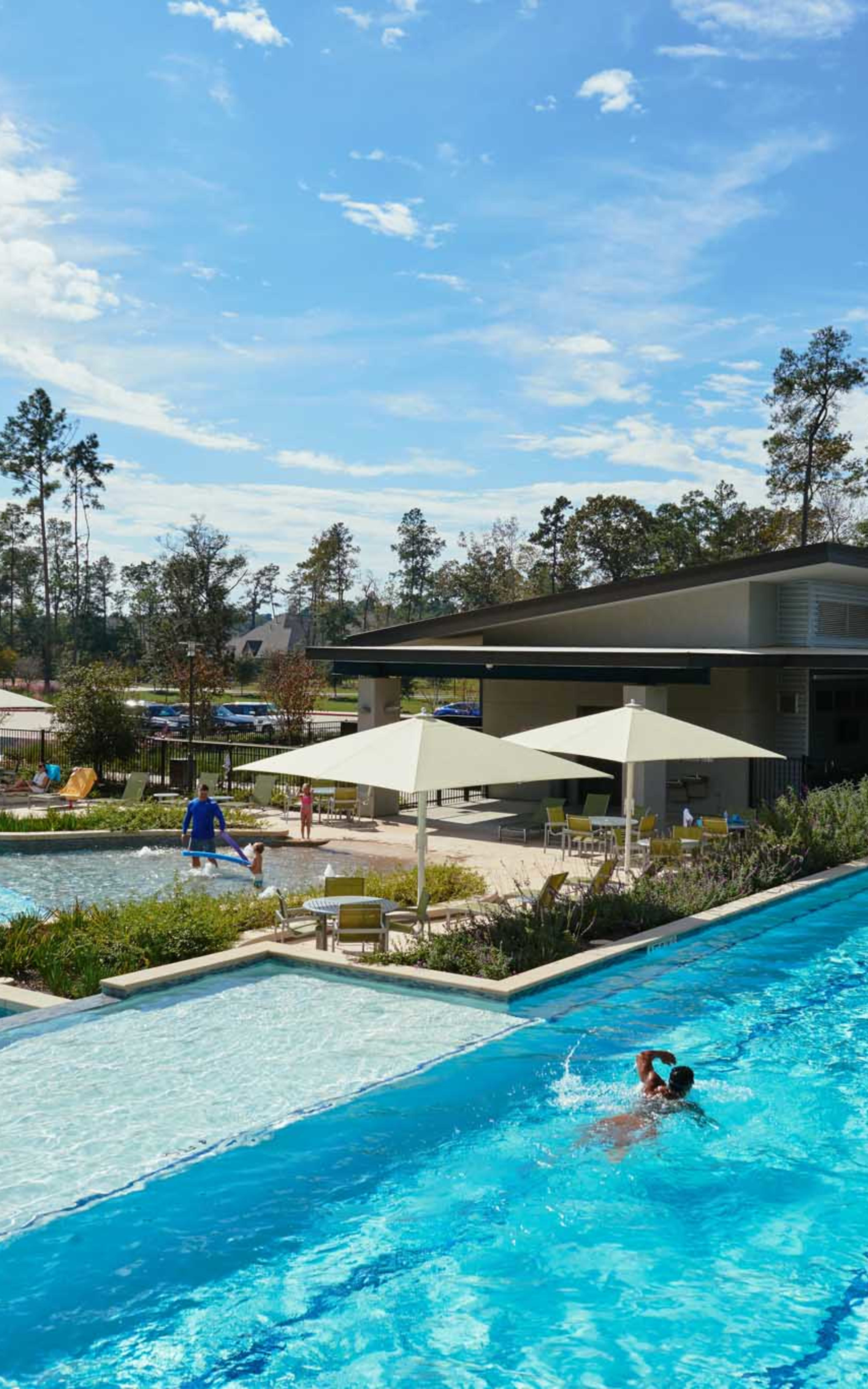 A large outdoor swimming pool surrounded by lush greenery, with a modern building and umbrellas in the background, creating a serene and inviting atmosphere.