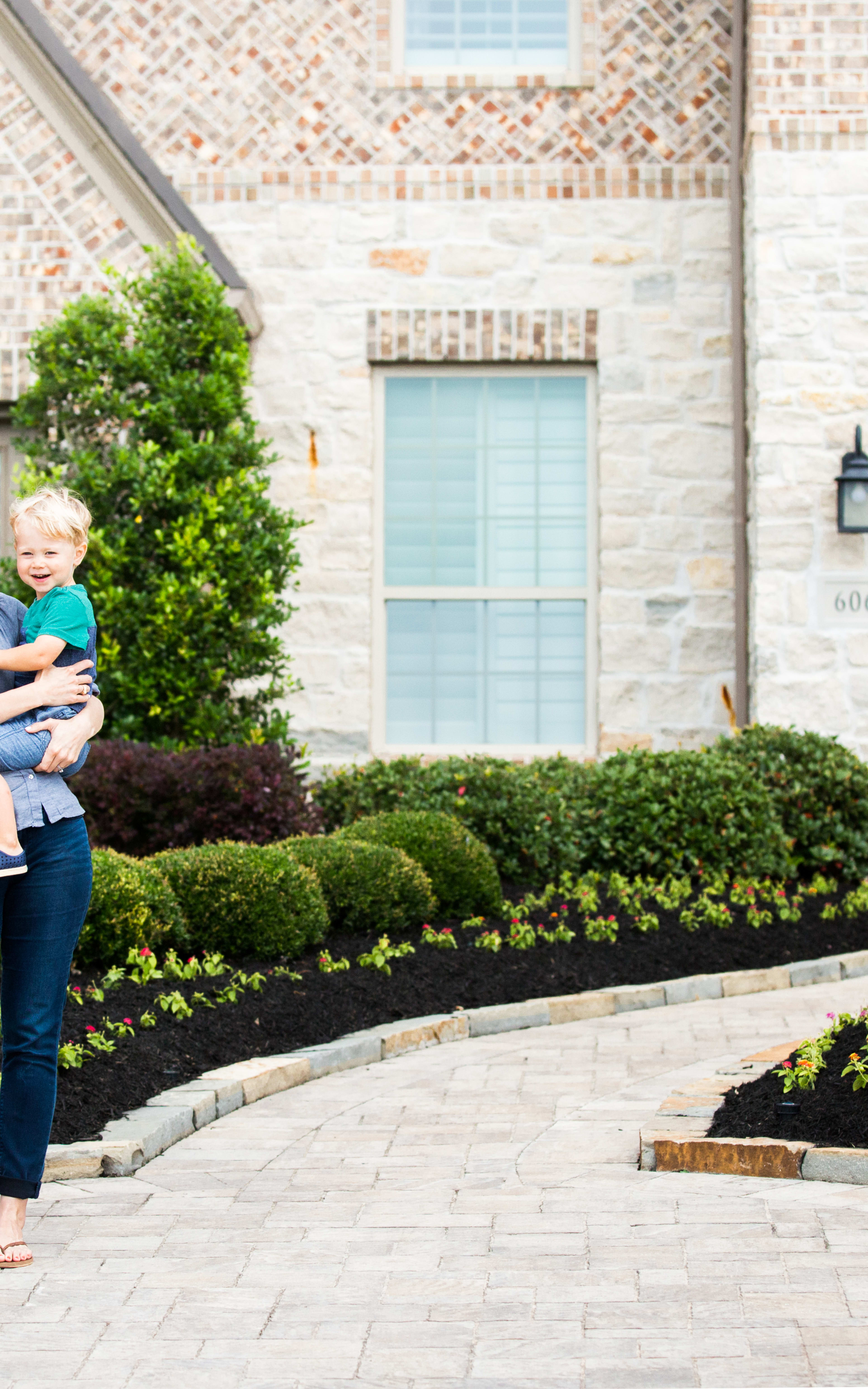 A woman holding a young child stands in front of a large, brick building with an arched entryway, surrounded by neatly trimmed bushes and a paved walkway.