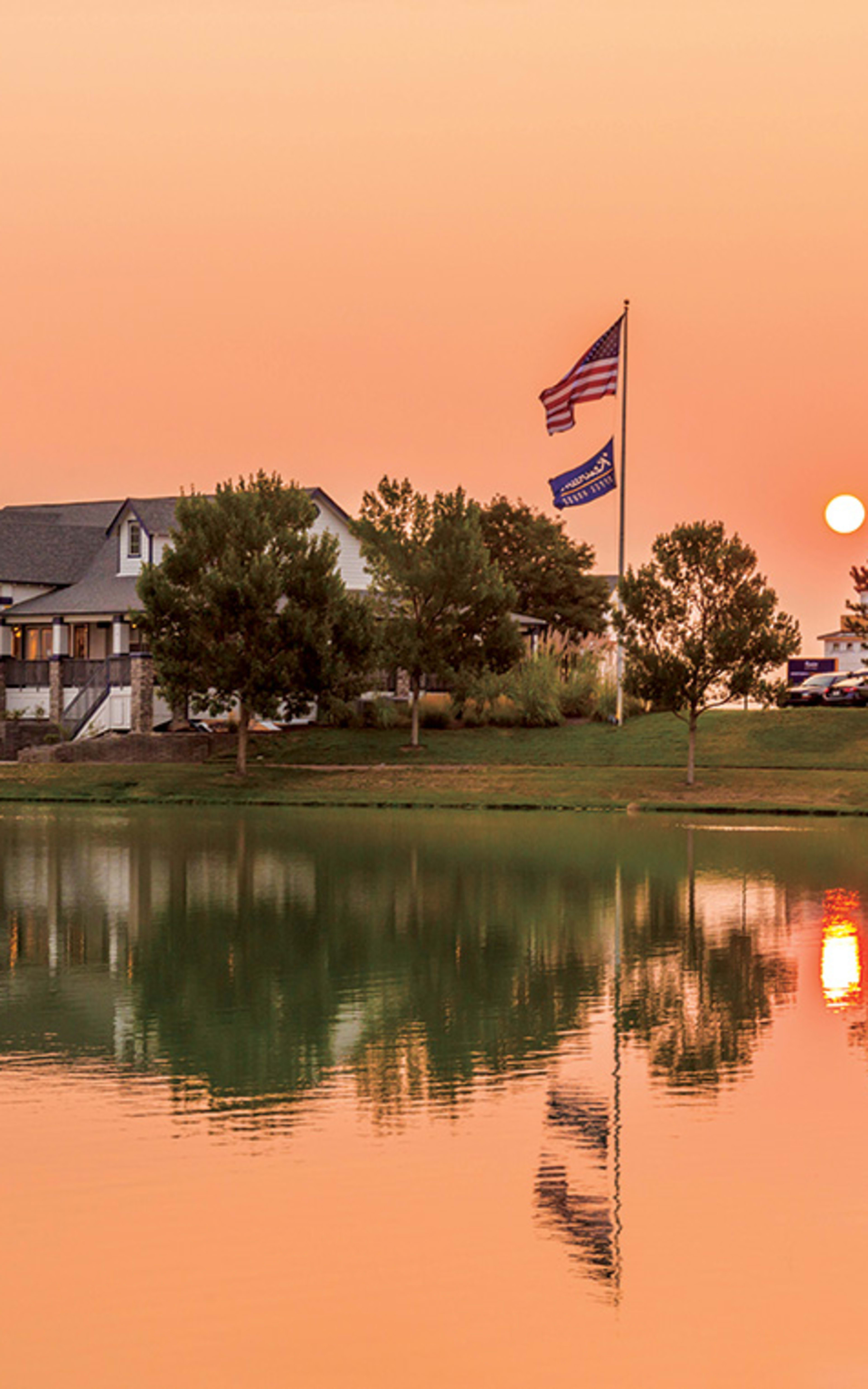 A picturesque scene of a large house with a flag-adorned roof reflected in a tranquil pond, surrounded by lush trees and a vibrant sunset sky.