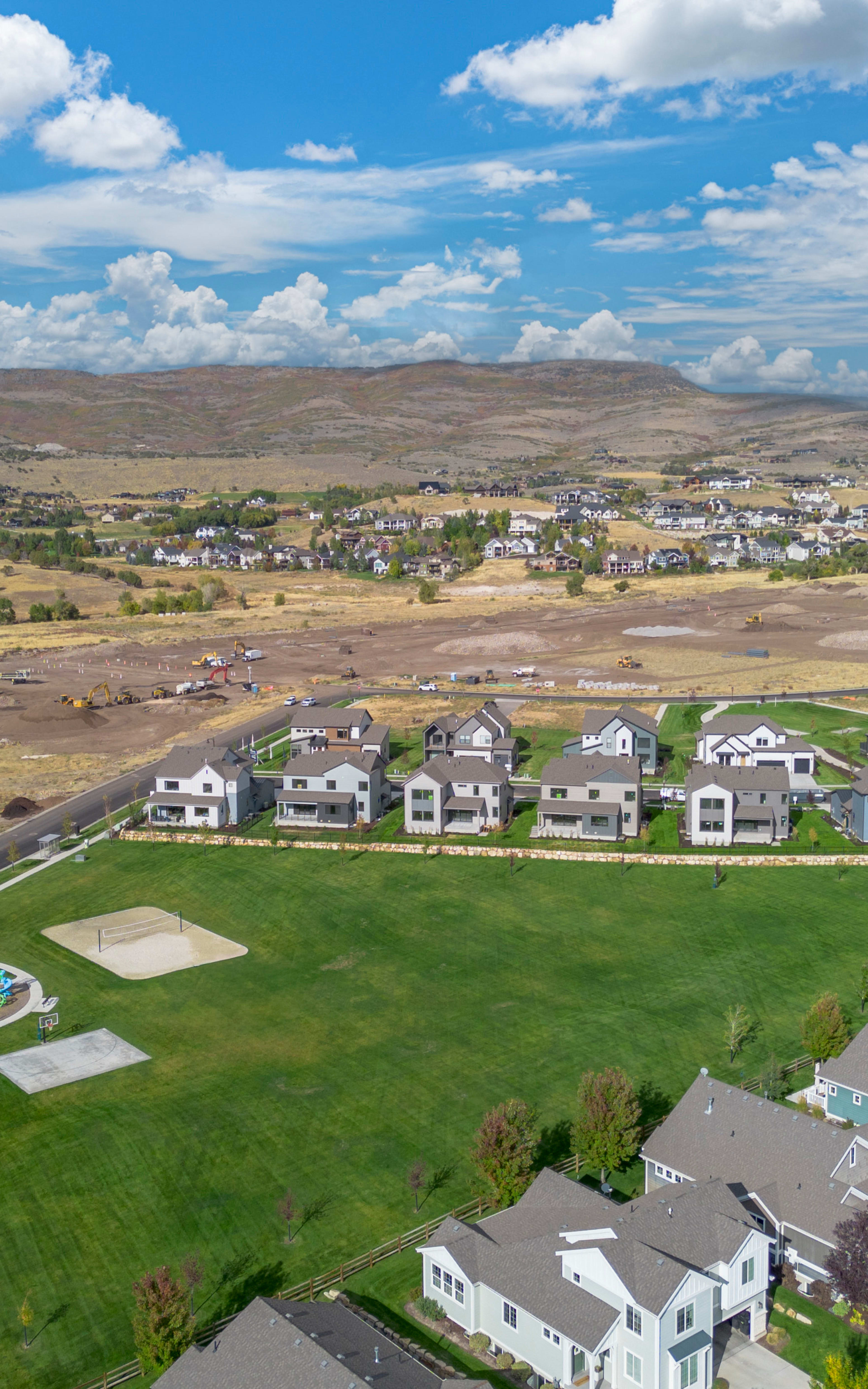 A sprawling suburban neighborhood nestled in a lush, rolling landscape, with a backdrop of distant hills and a vibrant blue sky dotted with fluffy white clouds.