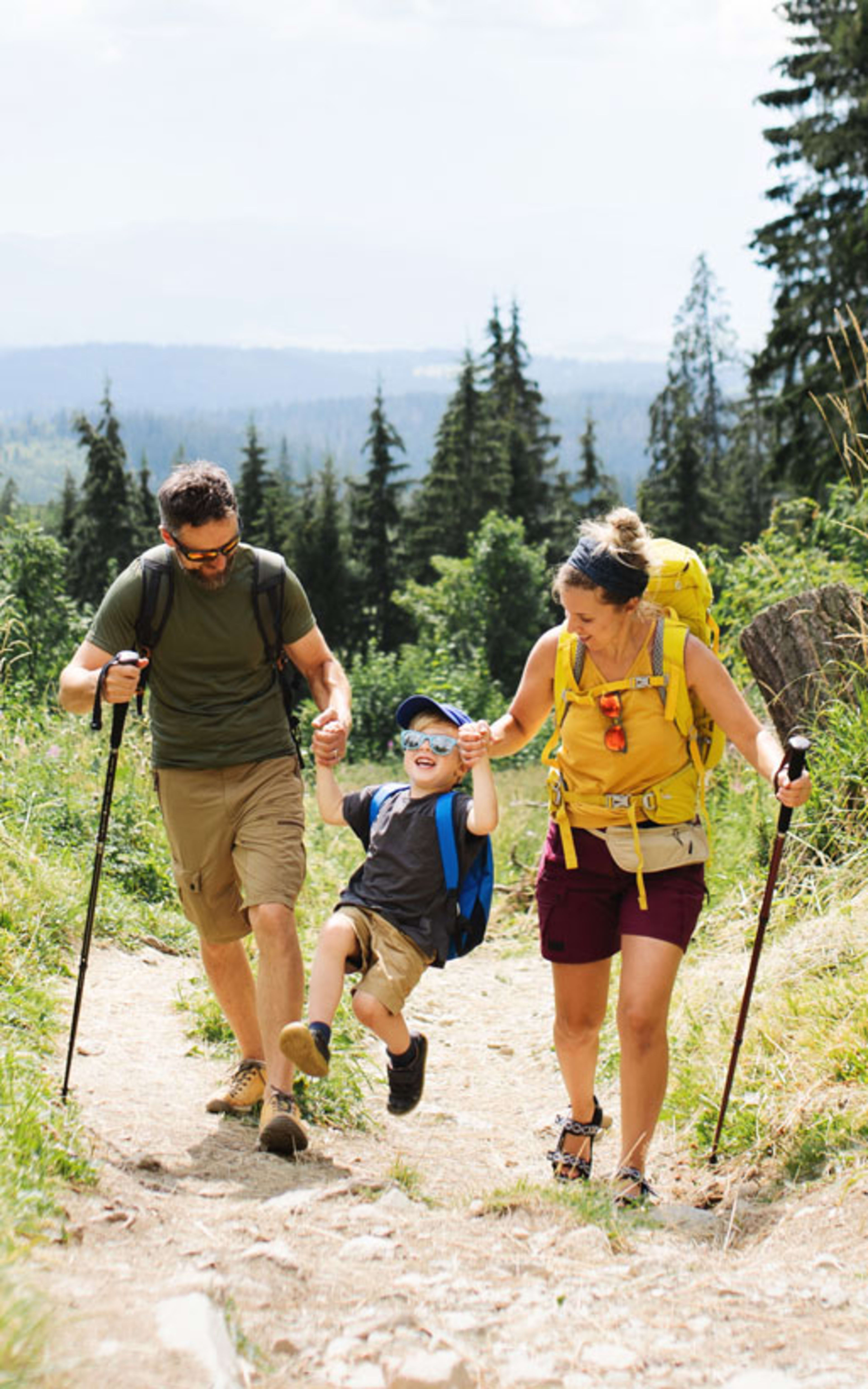 A family hiking together on a dirt trail through a lush, forested landscape with mountains visible in the distance.