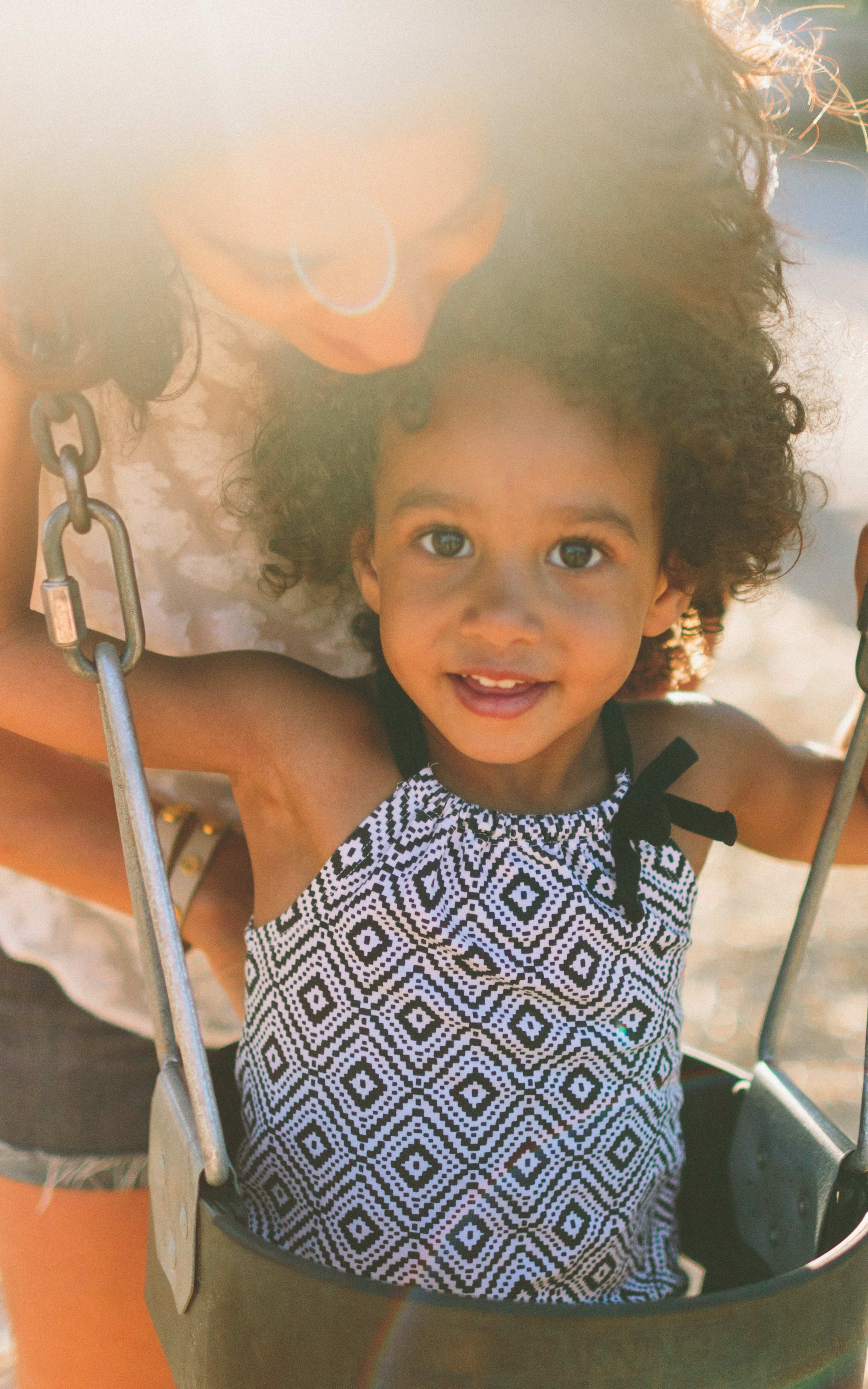 A young child with curly hair is happily swinging on a playground swing, surrounded by a bright and sunny outdoor environment.