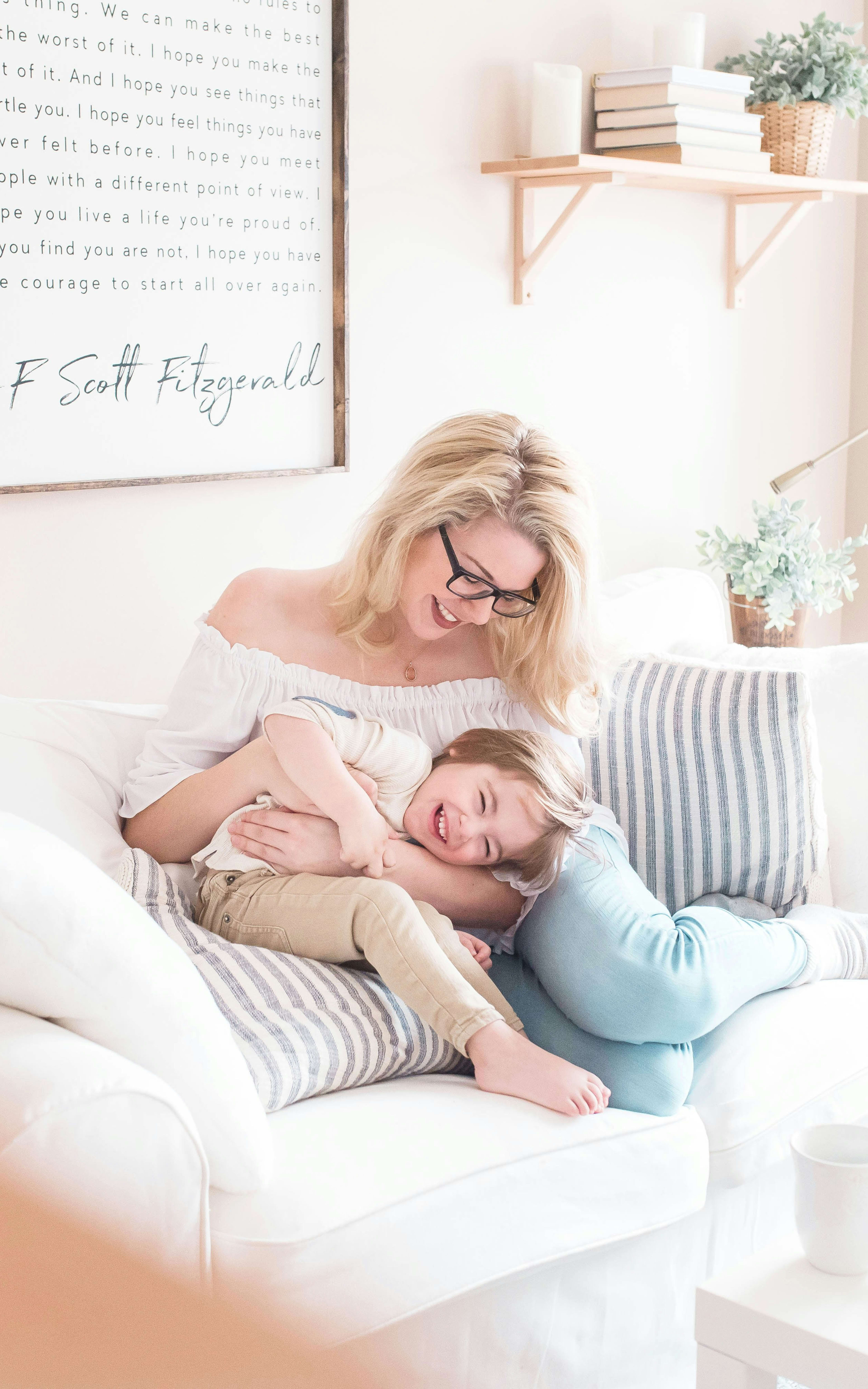 A woman is sitting on a couch, holding a sleeping baby in her arms, surrounded by a cozy and inviting home environment with shelves, decor, and natural light.