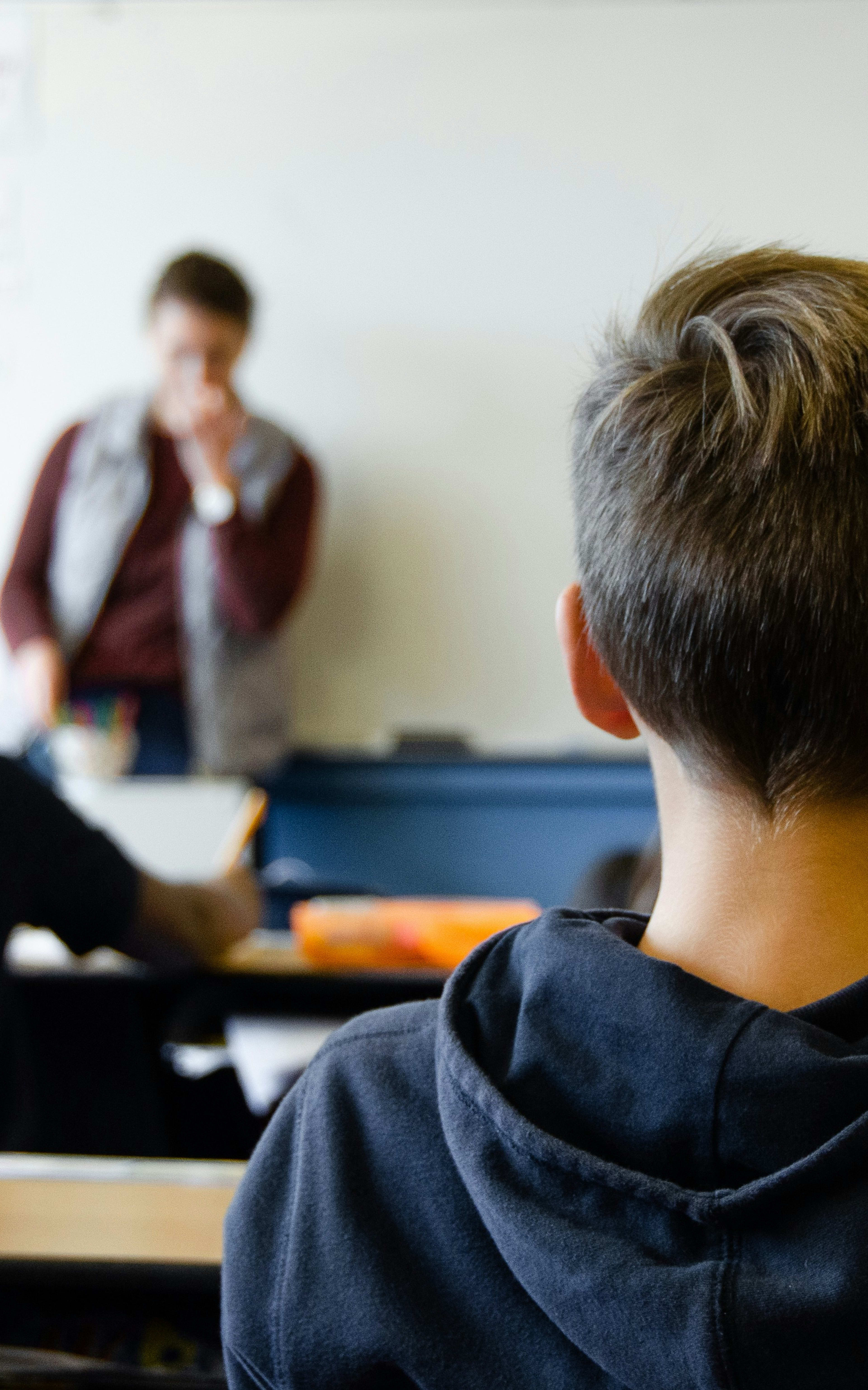 The image shows a classroom setting with students seated at desks, facing a teacher standing at the front of the room.