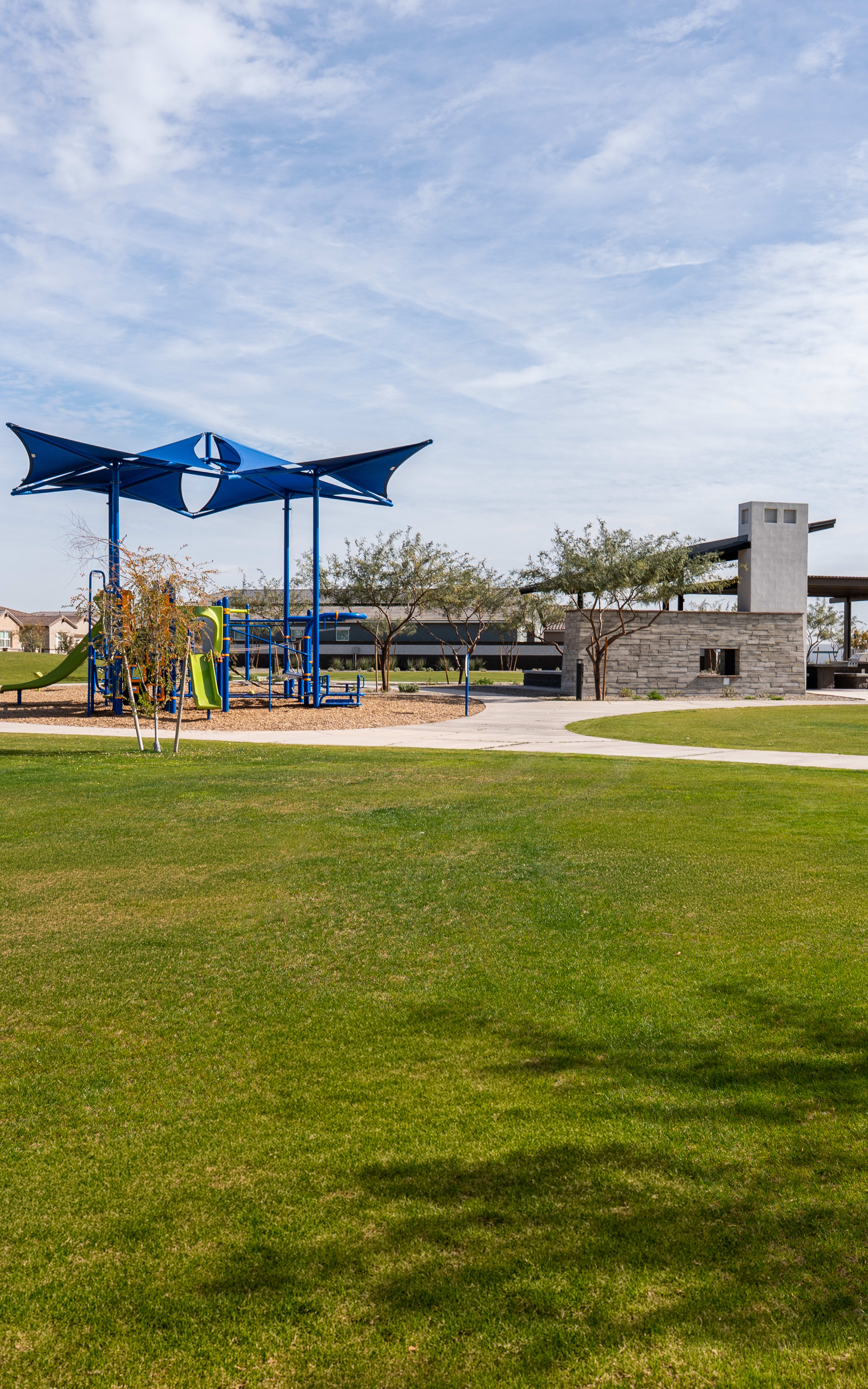 A grassy park with playground equipment, surrounded by residential buildings and a blue-and-white shade structure in the background.