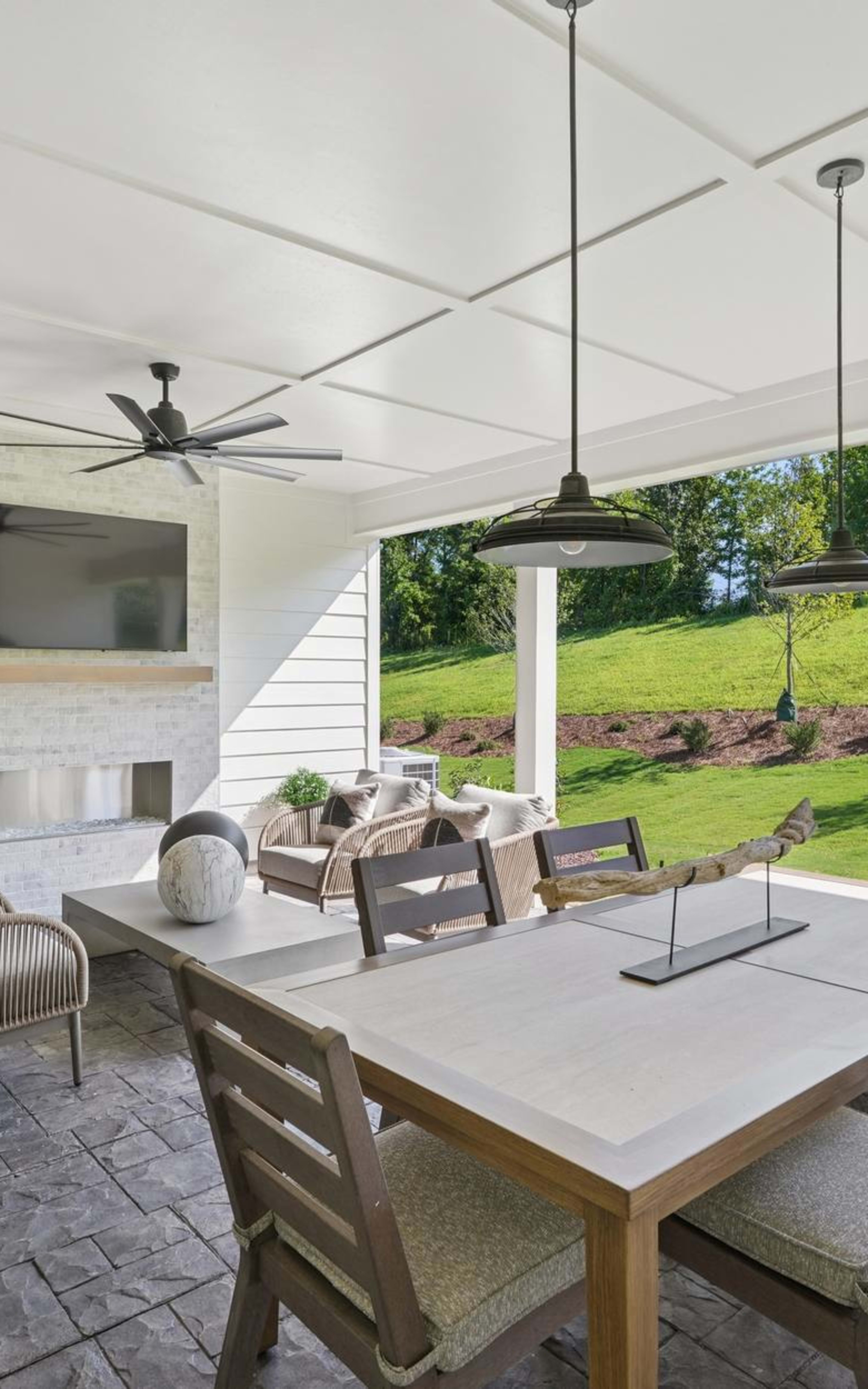 A cozy and inviting indoor-outdoor dining area with a wooden table, comfortable chairs, and a scenic view of a lush green lawn and trees through the large windows.