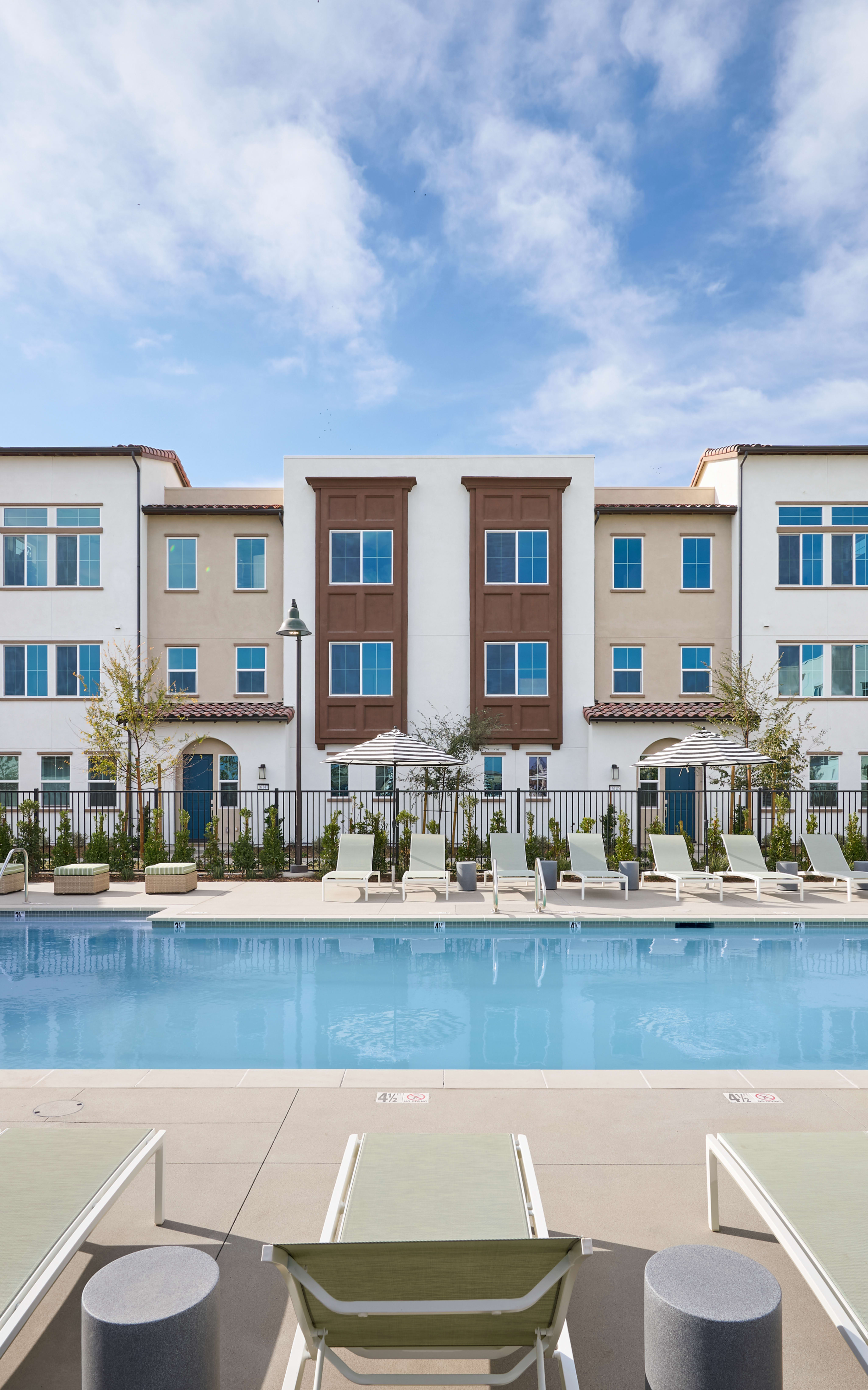 A modern multi-story apartment building with a swimming pool in the foreground, surrounded by lounge chairs and a lush outdoor environment under a bright, cloudy sky.