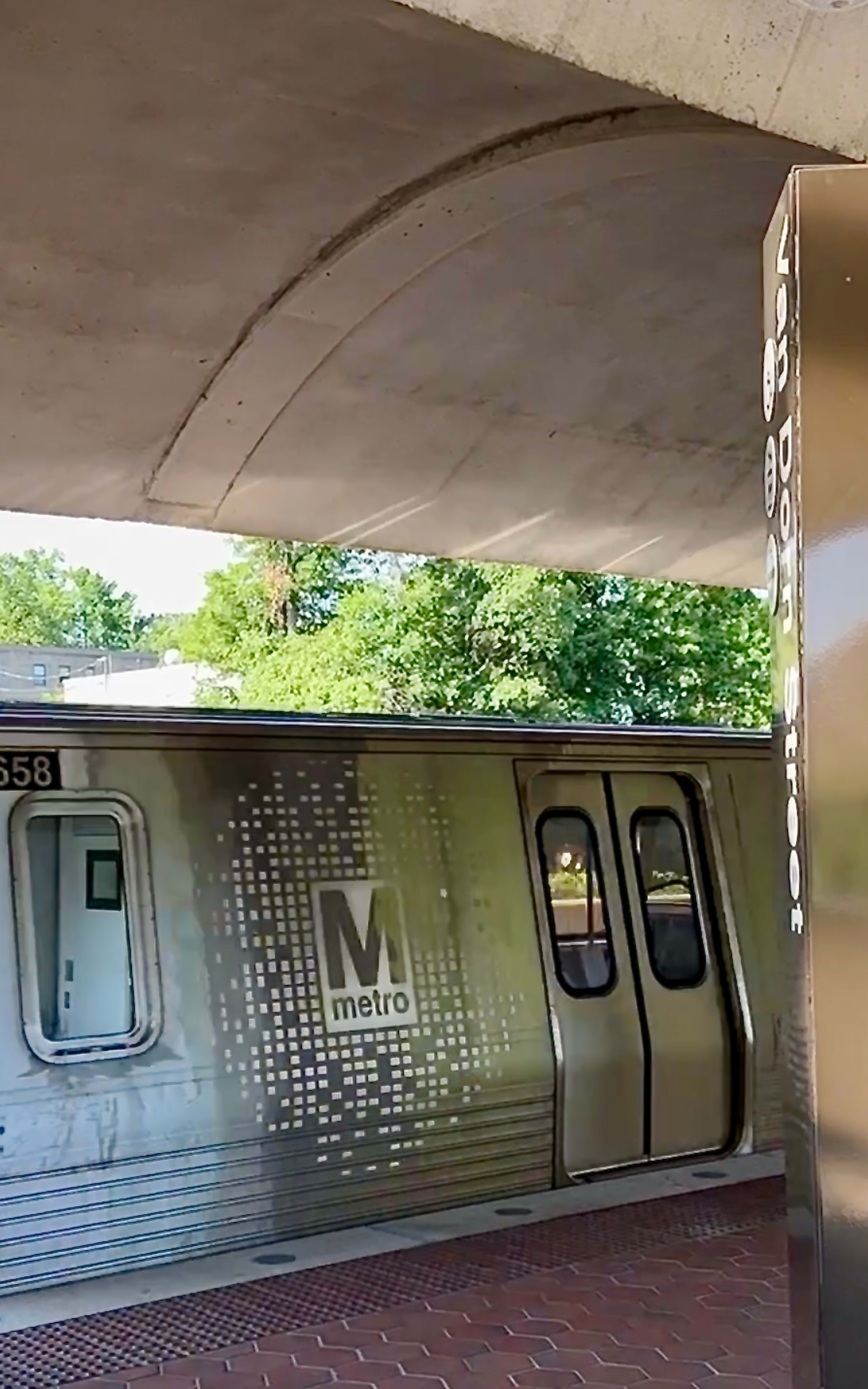 A subway train with the "Van Dorn Street" sign is parked under a concrete overpass, surrounded by greenery in the background.