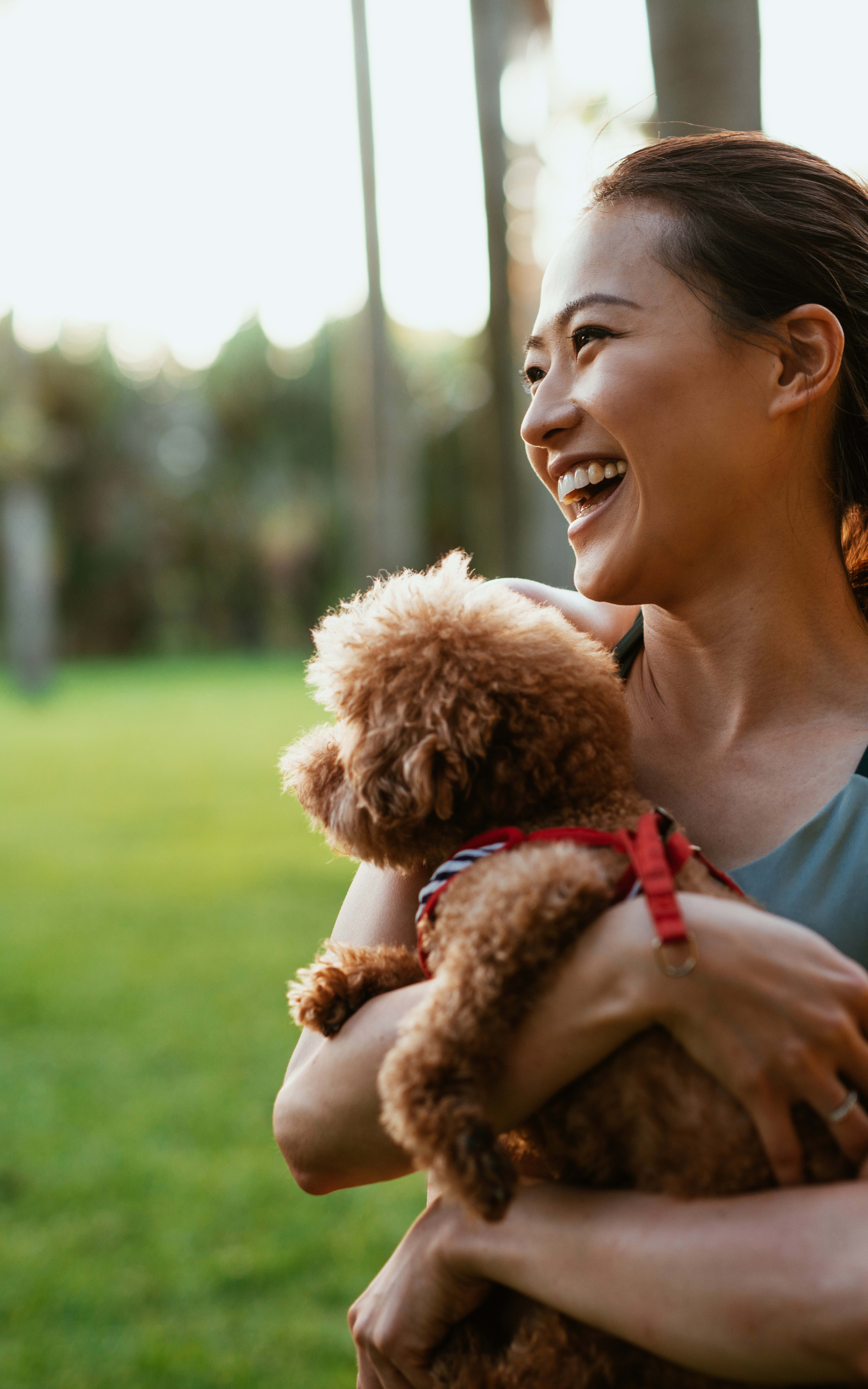 A smiling young woman is holding a small brown dog in a grassy park setting with trees in the background.