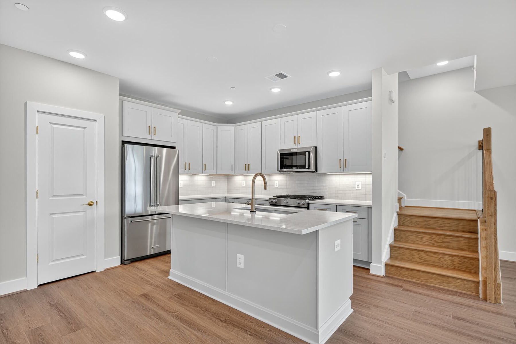 A modern, bright kitchen with white cabinets, stainless steel appliances, and hardwood floors, leading to a staircase in the background.