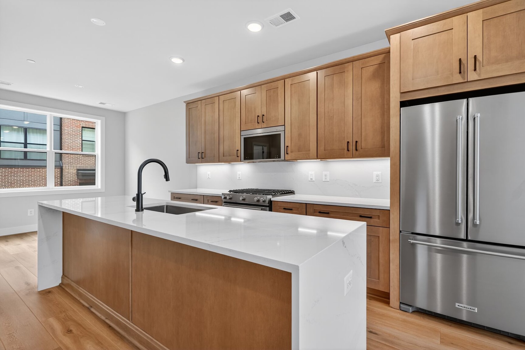 A modern kitchen with light wood cabinets, a white countertop, and stainless steel appliances, set against a backdrop of large windows providing natural light.