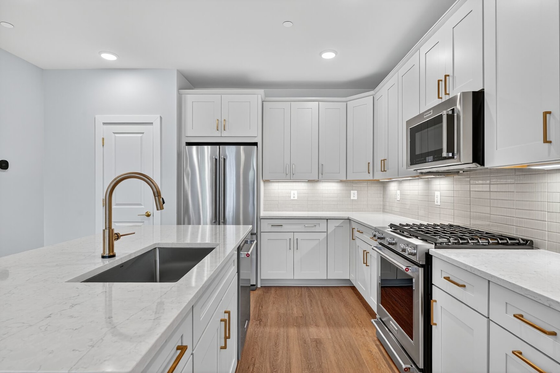 A modern, well-equipped kitchen with white cabinets, stainless steel appliances, and a marble countertop, set against a hardwood floor.