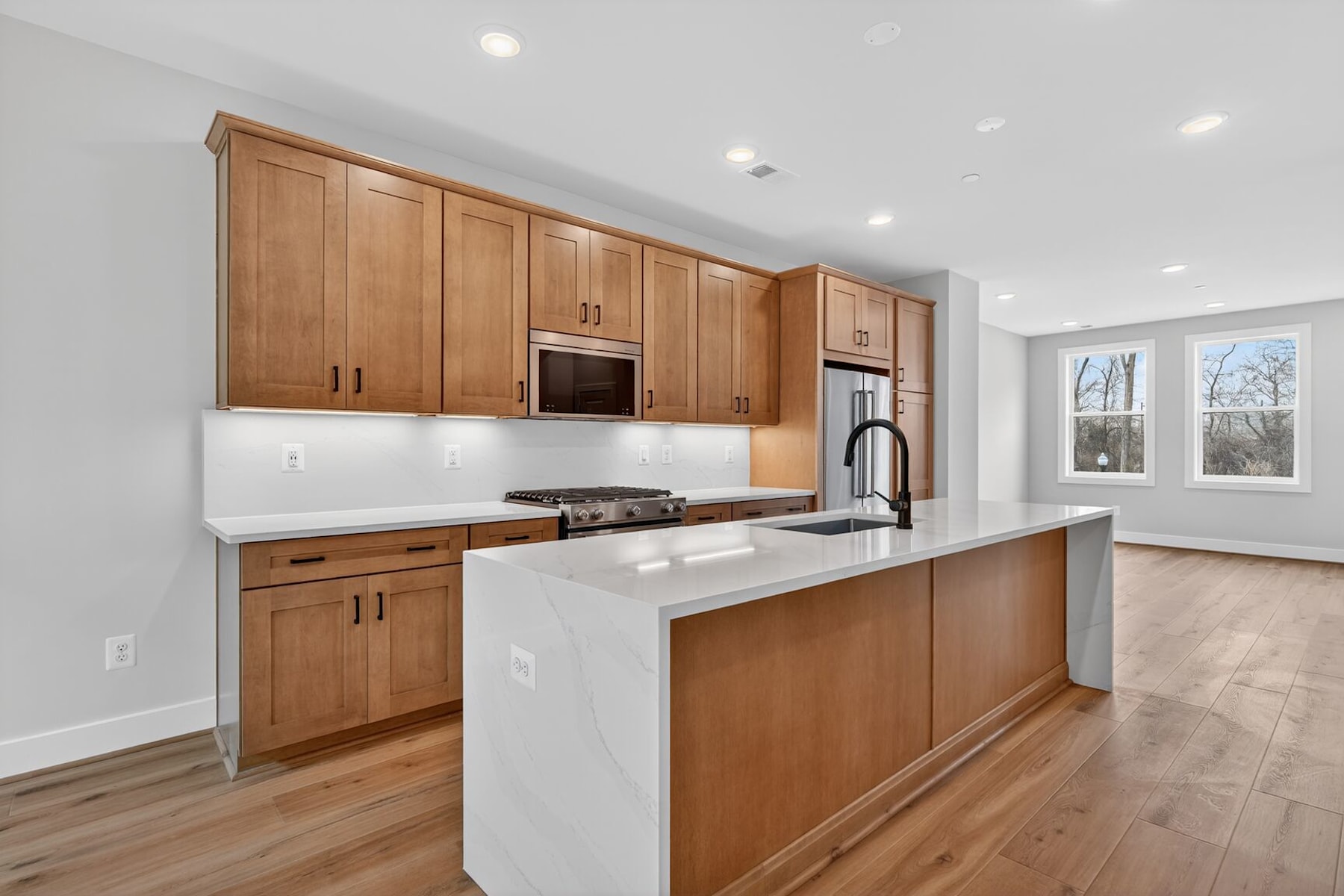 A modern kitchen with wooden cabinets, white countertops, and hardwood floors, set against a backdrop of large windows providing natural light.