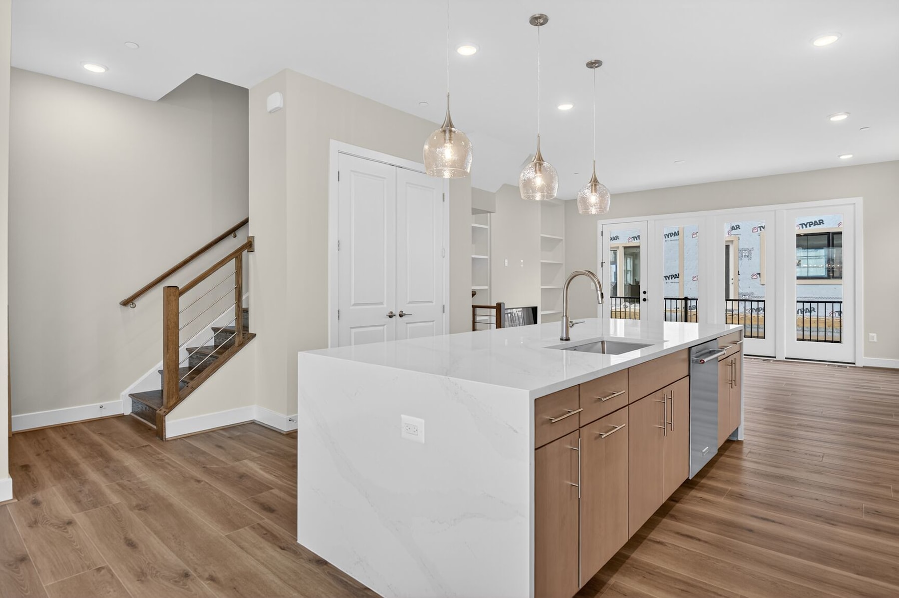 A modern, open-concept kitchen with white countertops, wooden cabinets, and pendant lighting fixtures, leading into a living area with a wooden staircase.