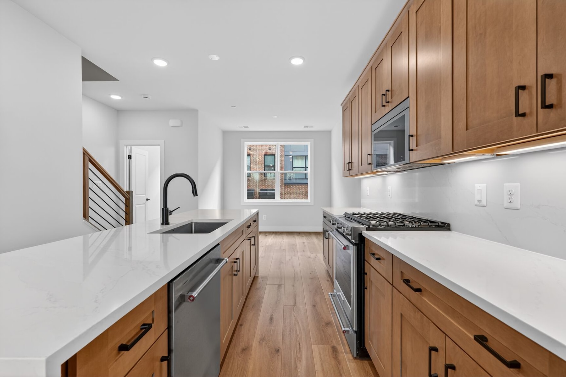 A modern, well-lit kitchen with wooden cabinets, white countertops, and stainless steel appliances, leading into a hallway with a door.