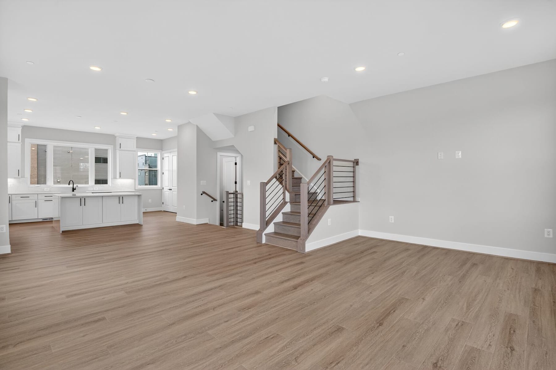 A spacious and modern interior with white walls, hardwood floors, and a staircase leading to an upper level. The room features a kitchen area with white cabinets and a central island.