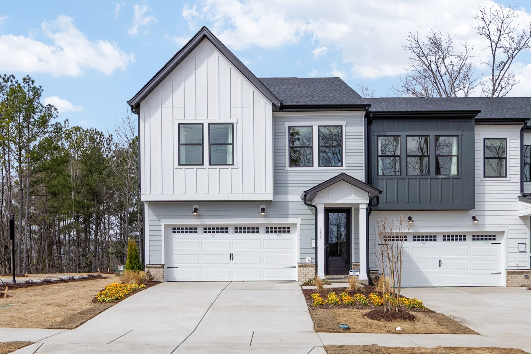 A modern two-story townhouse with a white exterior, black trim, and a garage door stands in a wooded area with trees in the background.