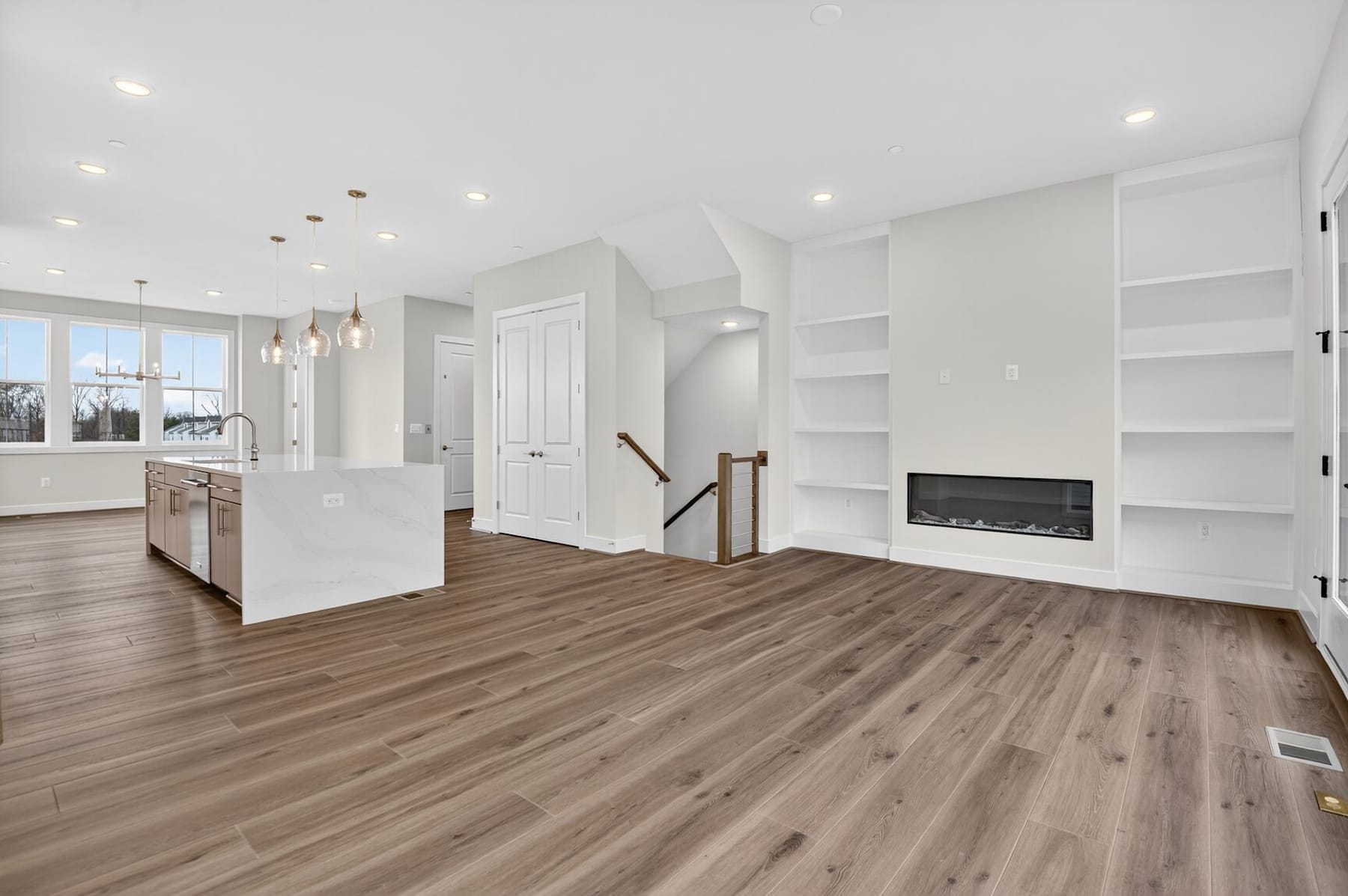 A spacious, modern living room with white walls, built-in shelves, and a fireplace, featuring a hardwood floor and large windows providing natural light.