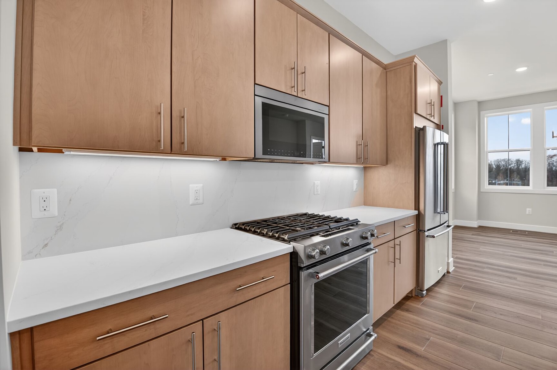 A modern kitchen with light wood cabinets, a stainless steel oven and refrigerator, and a white countertop.