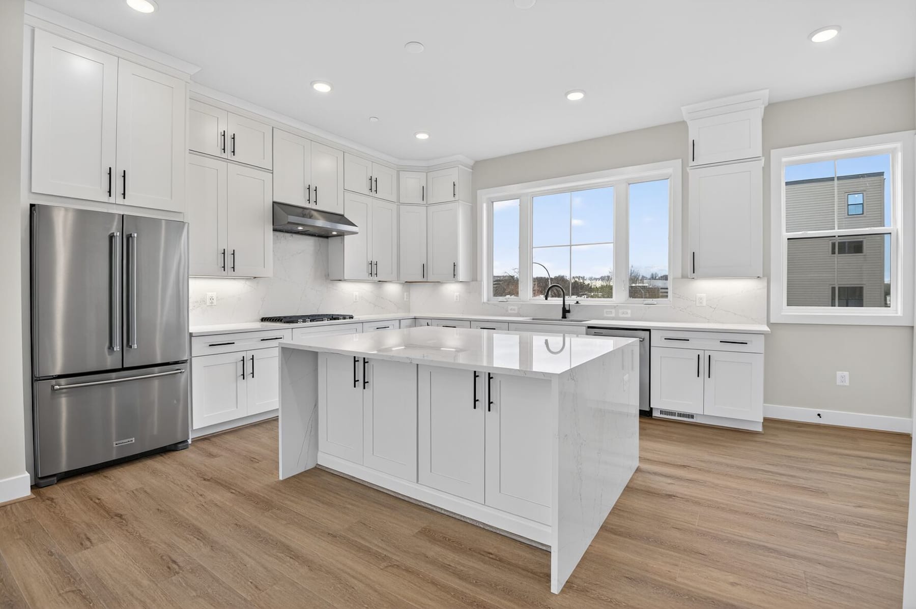 A modern, bright kitchen with white cabinets, stainless steel appliances, and a central island with a marble countertop, set against a backdrop of large windows overlooking an outdoor view.