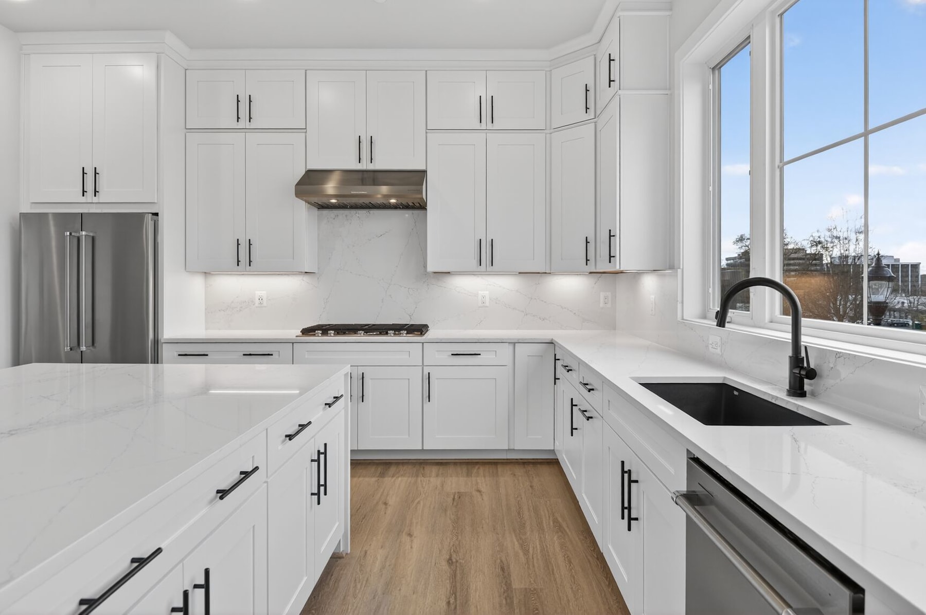 A modern, bright kitchen with white cabinets, stainless steel appliances, and a large window overlooking an outdoor view.