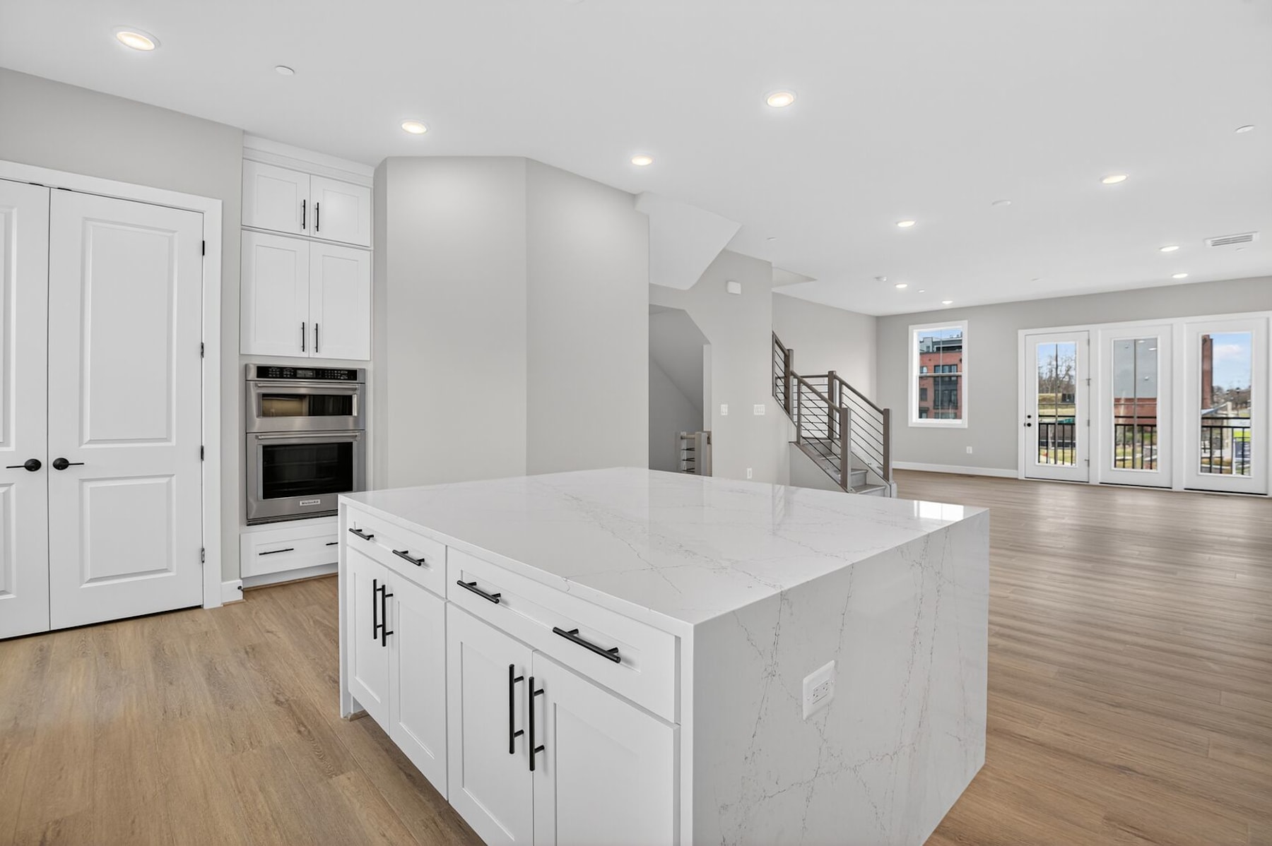 A modern, open-concept kitchen with white cabinets, a large island, and hardwood floors, with a staircase visible in the background.