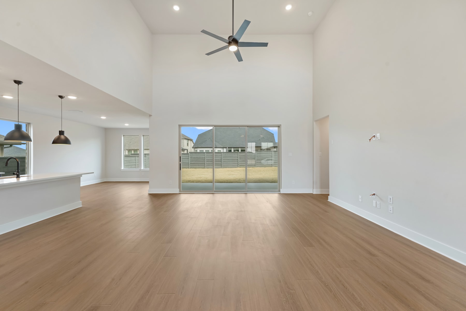 A spacious, open-concept living area with a high ceiling, hardwood floors, and a large ceiling fan. The room features a sliding glass door leading to an outdoor area, providing natural light and a connection to the exterior.