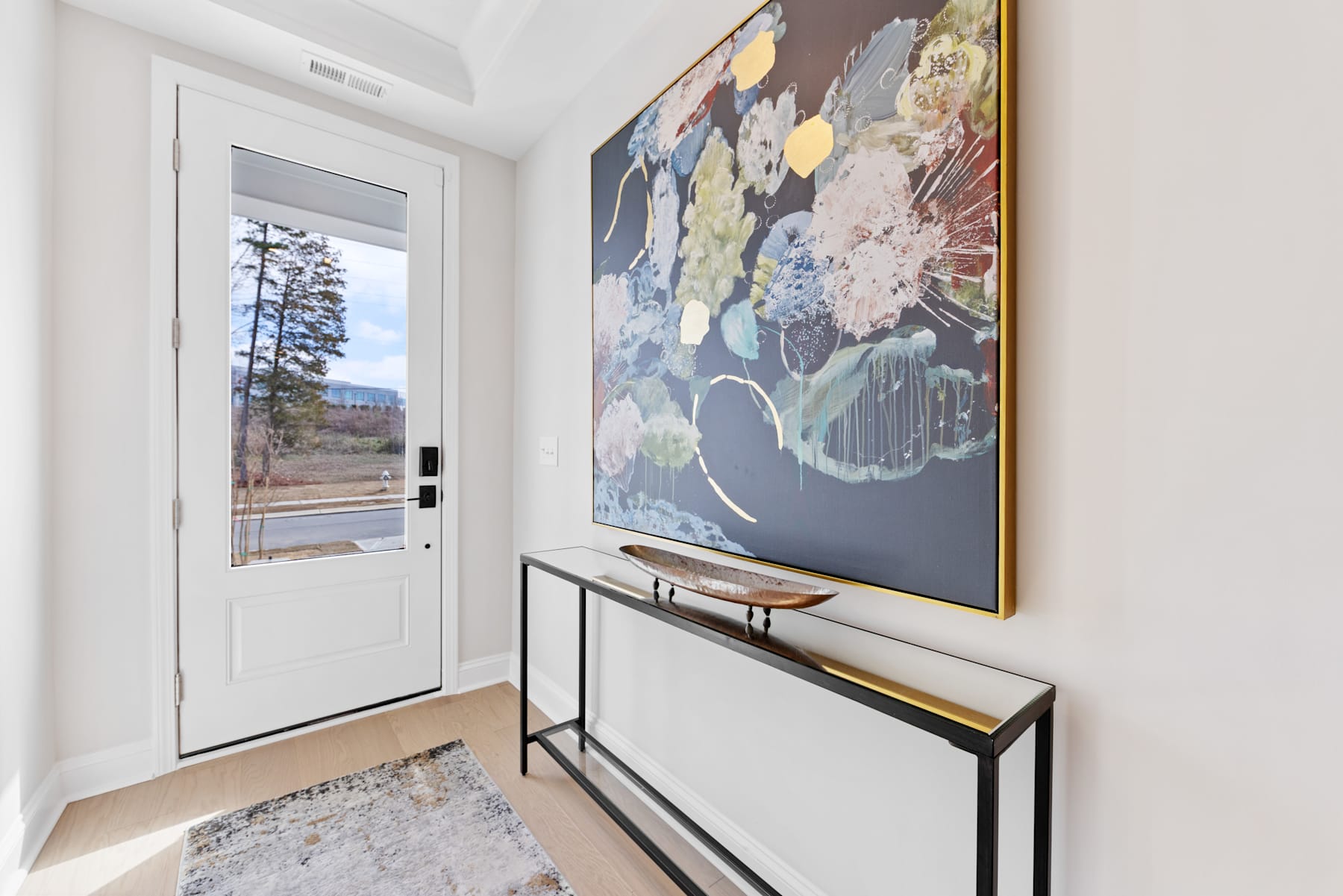 A modern and minimalist entryway with a large abstract artwork on the wall, a sleek console table, and a glass door providing a view of the outdoor scenery.
