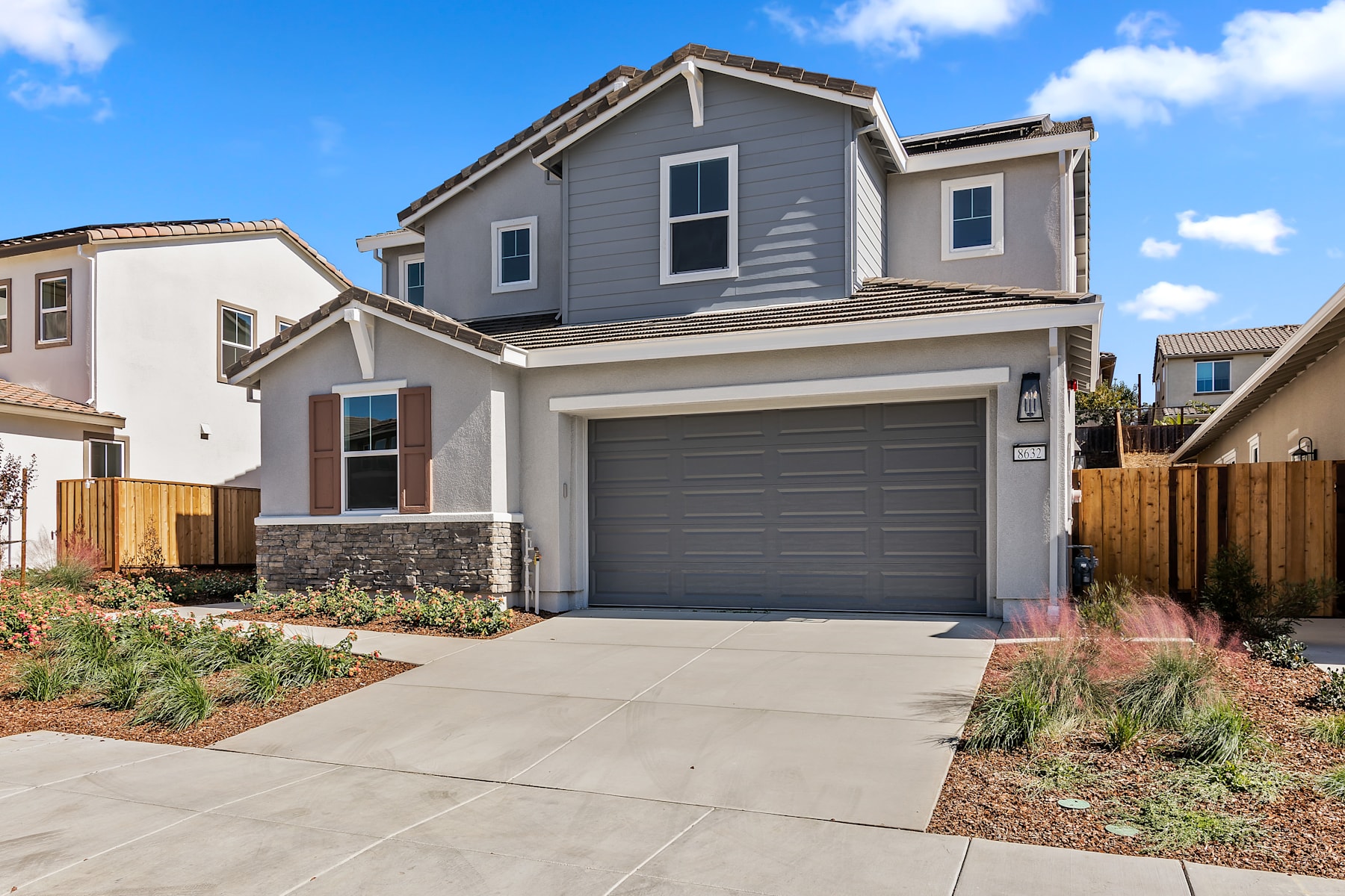 A two-story residential house with a gray exterior, a garage door, and a paved driveway in the foreground, set against a backdrop of a blue sky with scattered clouds.