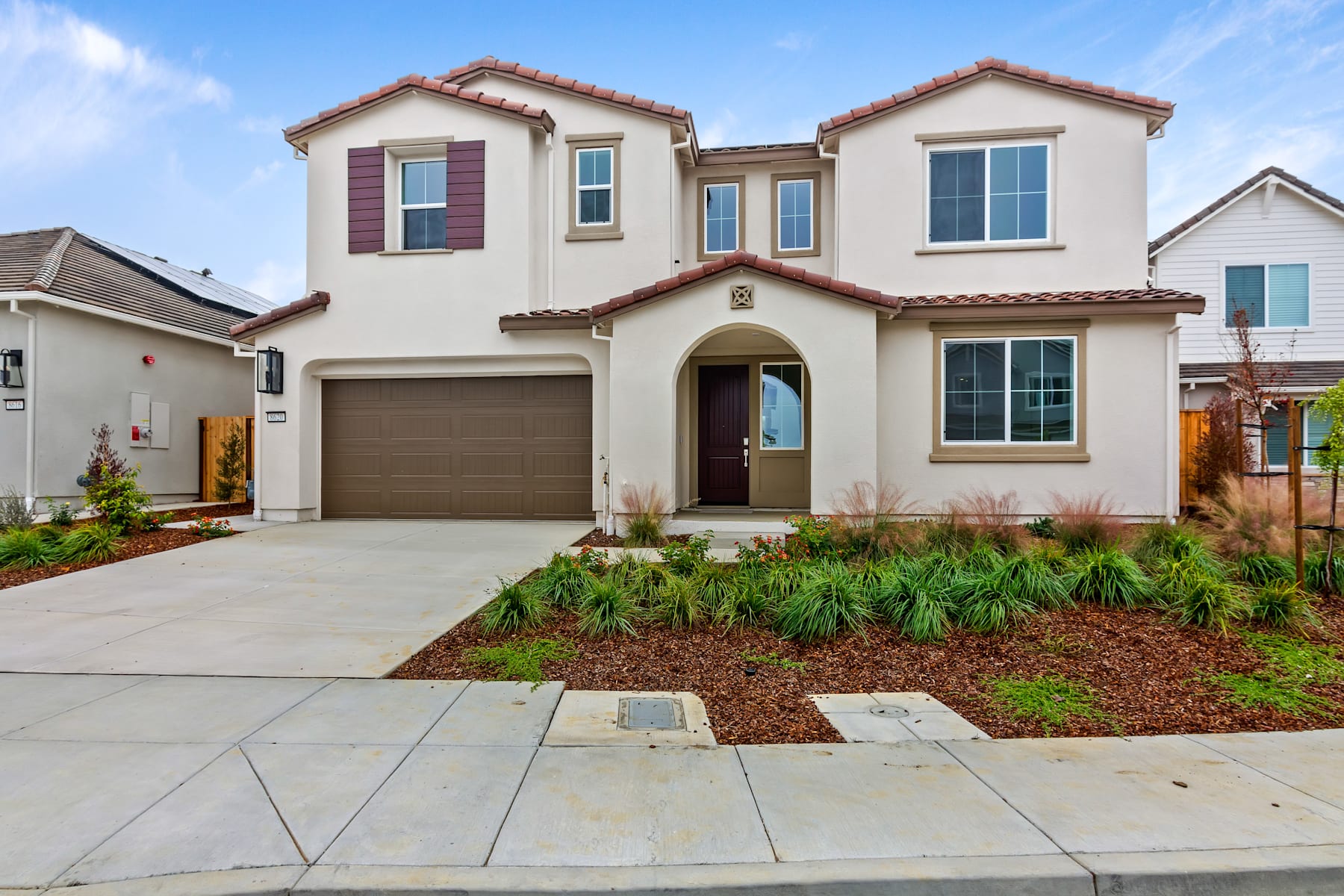 A two-story Mediterranean-style house with a red tile roof, surrounded by a well-landscaped yard featuring a paved walkway, shrubs, and a grassy area.