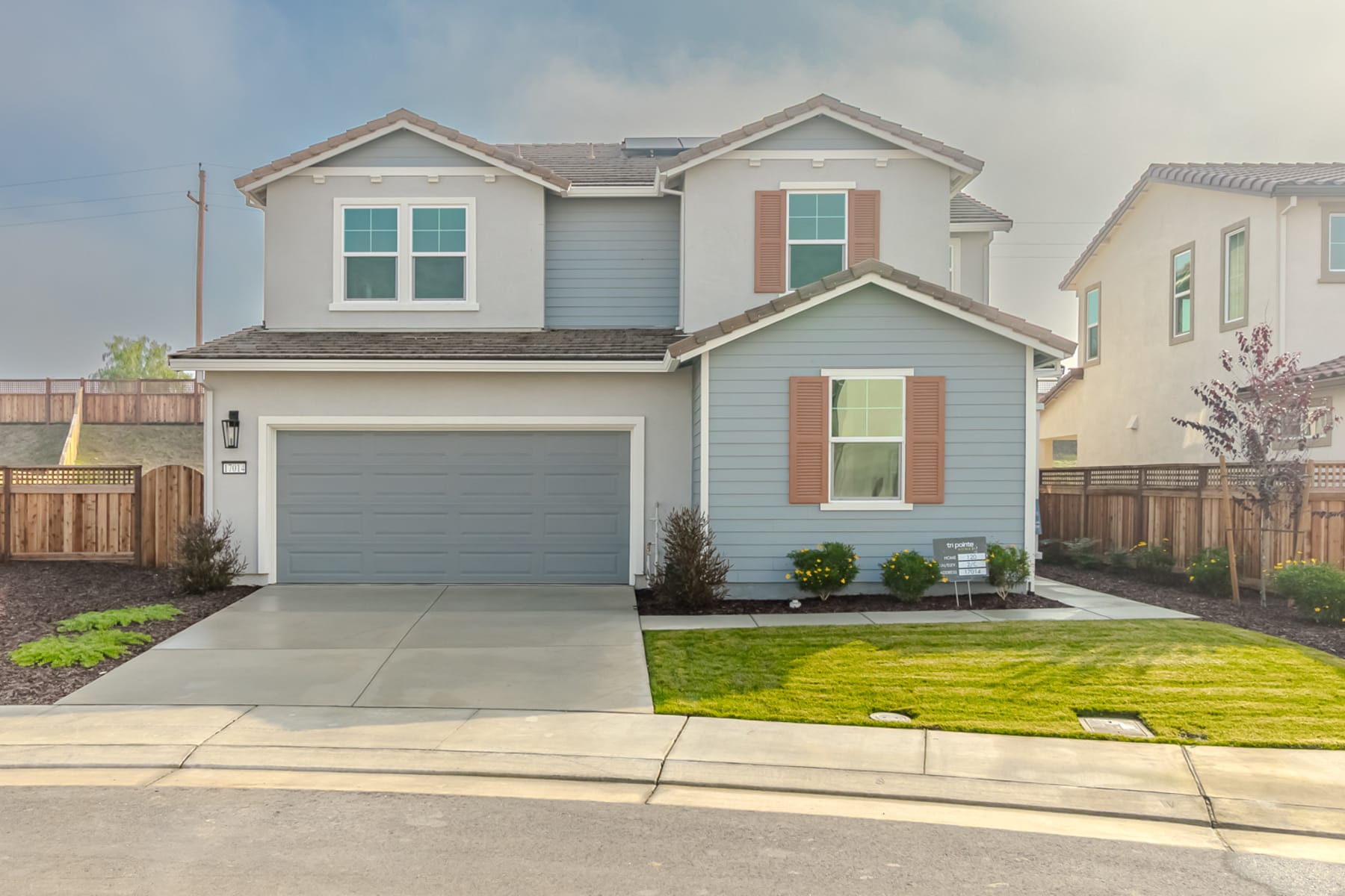 A two-story residential house with a well-manicured lawn, surrounded by a paved driveway and a wooden fence in the foreground, set against a clear blue sky in the background.